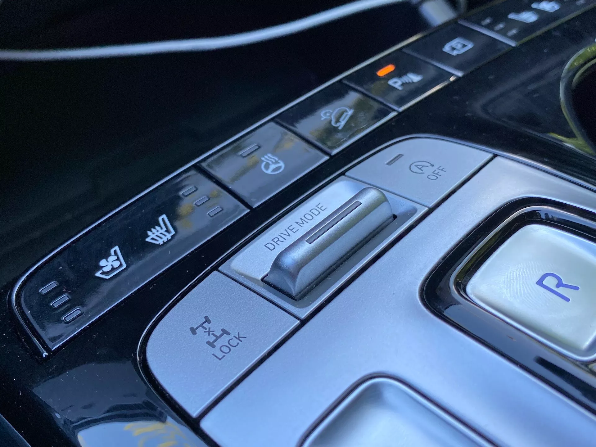 Vista ravvicinata della consolle centrale della Hyundai Tucson 2020, con particolare attenzione al selettore della modalità di guida e ai vari pulsanti di controllo. L'immagine rivela un avanzato interruttore di modalità di guida per diverse condizioni di guida, un pannello per il controllo della trazione, un pulsante per la retromarcia e ulteriori controlli per la stabilità del veicolo e le funzioni di assistenza al parcheggio. Il design elegante e i materiali high-tech sottolineano l'interno automobilistico sofisticato di questa auto moderna di lusso.