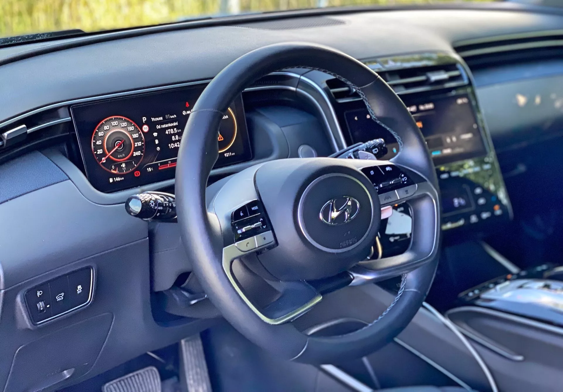 Vista interna di una Hyundai Tucson 2020 dalla prospettiva del conducente che mostra il volante moderno con comandi multifunzionali e un quadro strumenti digitale che mostra il tachimetro, il contagiri, il livello del carburante e la temperatura del motore. L'immagine evidenzia anche una parte della console centrale con un grande sistema di infotainment touchscreen, un design pulito del cruscotto con illuminazione ambientale e materiali di alta qualità che suggeriscono finiture di lusso.