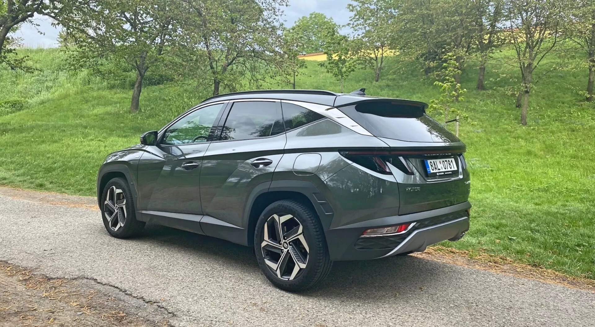 Prospettiva posteriore da tre quarti di un SUV Hyundai Tucson 1.6 T-GDi Pure del 2020 parcheggiato su una strada stretta in un contesto rurale, che mostra grandi cerchi in lega multirazze, vernice metallizzata scura con una linea del tetto discendente, spoiler posteriore, vetri posteriori oscurati, luci posteriori moderne e doppi terminali di scarico integrati nel paraurti posteriore.
