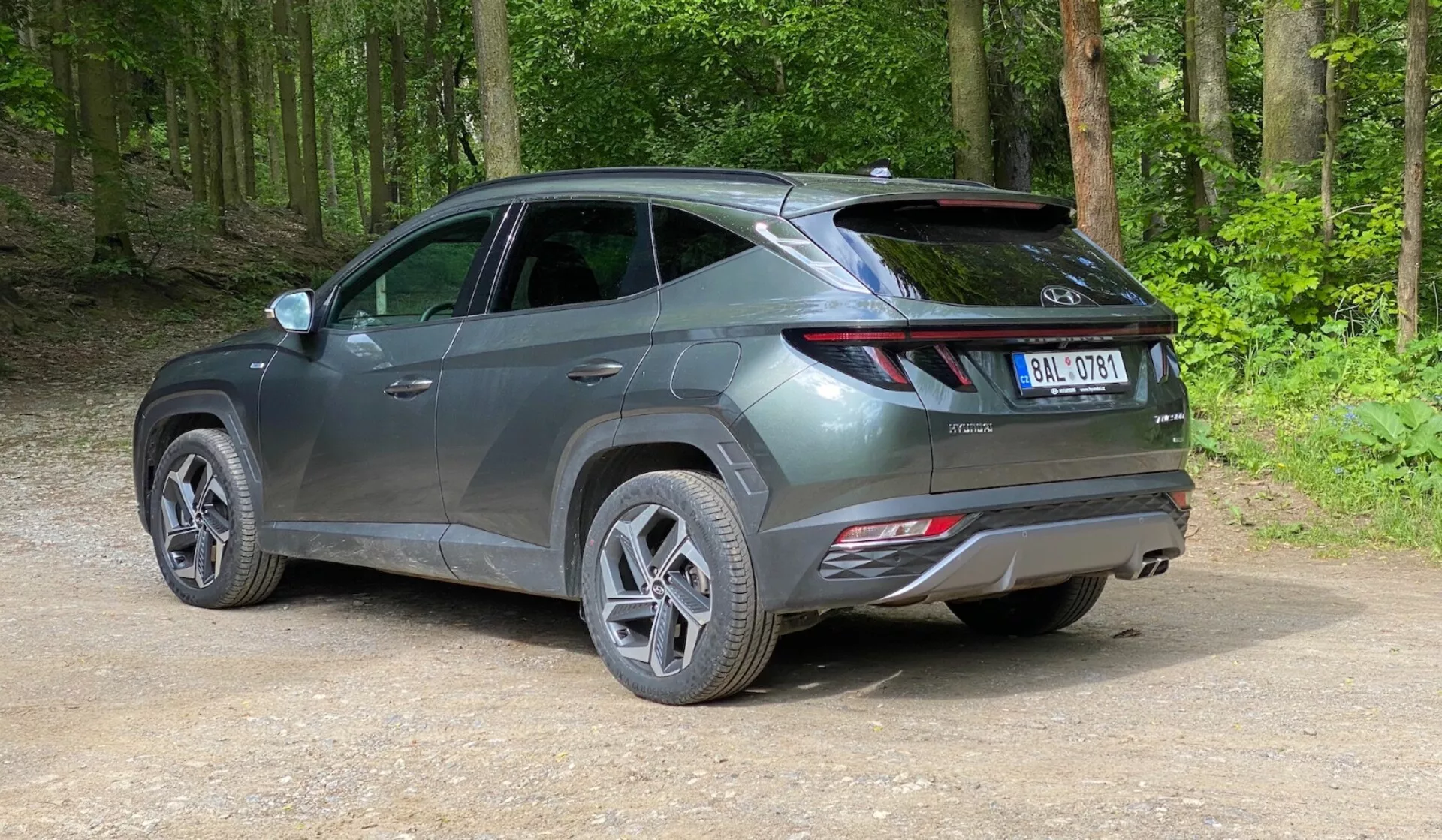 Un SUV moderno parcheggiato su un sentiero non asfaltato in un'area forestale, catturato da una vista posteriore a tre quarti da una leggera elevazione. Il veicolo presenta un design elegante e contemporaneo con linee affilate, luci posteriori allungate che si avvolgono dalla parte posteriore ai lati, uno spoiler visibile e riflettori sul paraurti posteriore, suggerendo caratteristiche di sicurezza avanzate. Il veicolo in grigio metallizzato ha cerchi in lega multiraggio e pneumatici stradali, con una superficie lucida che indica che è nuovo o ben mantenuto. Intorno all'auto c'è una foresta verde e rigogliosa con terra battuta e ghiaia sciolta sul terreno.