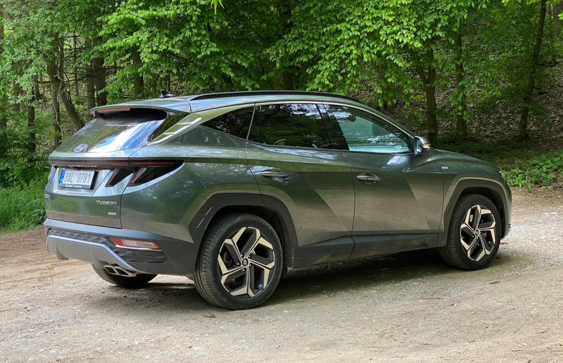 Vista angolare posteriore tre quarti di un moderno SUV Hyundai Tucson 2020 con un focus sul profilo laterale e il design posteriore. Il veicolo è posizionato sulla sinistra con la foto scattata da destra, enfatizzando l'angolo posteriore destro. Le luci posteriori a LED distintive avvolgono dal retro verso i lati e uno spoiler montato sul tetto aggiunge un tocco sportivo. La linea di cintura ascendente, il profilo del tetto inclinato, i finestrini oscurati e le maniglie delle portiere in tinta con la carrozzeria enfatizzano il suo aspetto da coupé. I grandi cerchi in lega con design multi-razza e i pneumatici a profilo ribassato migliorano il suo aspetto lussuoso. Dettagli minori come il tergilunotto e le modanature degli archi ruota indicano praticità e robustezza. L'auto è posizionata sulla ghiaia in un ambiente naturale con alberi sullo sfondo.