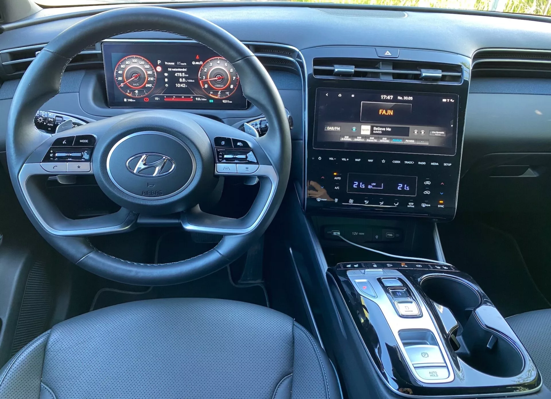 Vista interna della Hyundai Tucson 2020 dal sedile del conducente, che mostra il volante con logo Hyundai e comandi per audio, telefono e impostazioni del cruise control. Il cruscotto presenta un pannello strumenti digitale con tachimetro e contagiri accennati in rosso. La console centrale ospita un sistema di infotainment con touchscreen che mostra i controlli dell'aria condizionata, pulsanti fisici e manopole per il controllo del clima, e un cambio automatico con selettore elettronico. È visibile una possibile base di ricarica wireless, rivestimenti in tessuto scuro e una cabina ben illuminata.