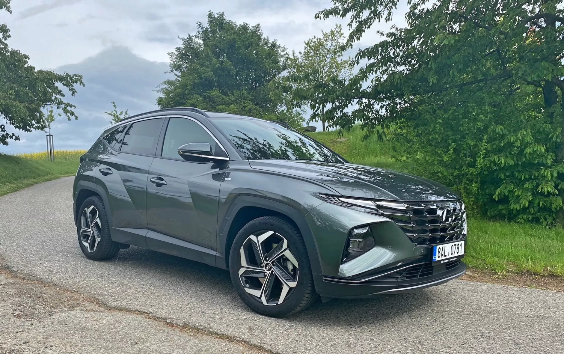 L'immagine mostra un moderno SUV Hyundai Tucson 1.6 T-GDi Pure parcheggiato su una strada asfaltata con uno sfondo naturale. Fotografato da un'angolazione frontale di tre quarti, la griglia prominente, i fari eleganti, il paraurti anteriore con fendinebbia o prese d'aria, le grandi ruote in lega con design multiraggio e gli specchietti laterali sono tutti visibili. La silhouette del SUV mostra una linea del tetto che si inclina verso la parte posteriore, indicativa di un design sportivo e aerodinamico. Catturata in condizioni di luce diurna soffusa, la foto mette in risalto il colore e i dettagli del veicolo senza ombre forti.