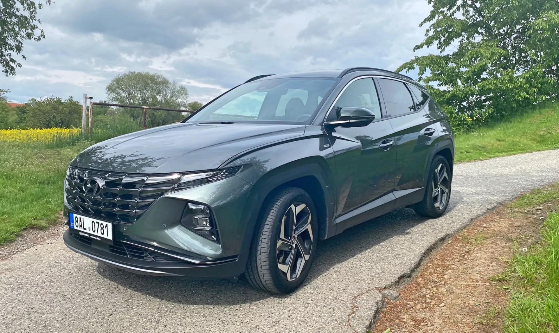 Hyundai Tucson 2020 1.6 T-GDI Pure SUV, visto da un'angolazione frontale di tre quarti su una strada asfaltata con alberi ed erba sullo sfondo. L'immagine mostra la griglia del veicolo, i fari a LED affilati, i cerchi in lega, gli specchietti laterali con indicatore di direzione integrato e la vernice metallizzata scura. Sono visibili le porte anteriori del conducente e posteriori. Foto scattata durante il giorno in condizioni di luce nuvolosa.