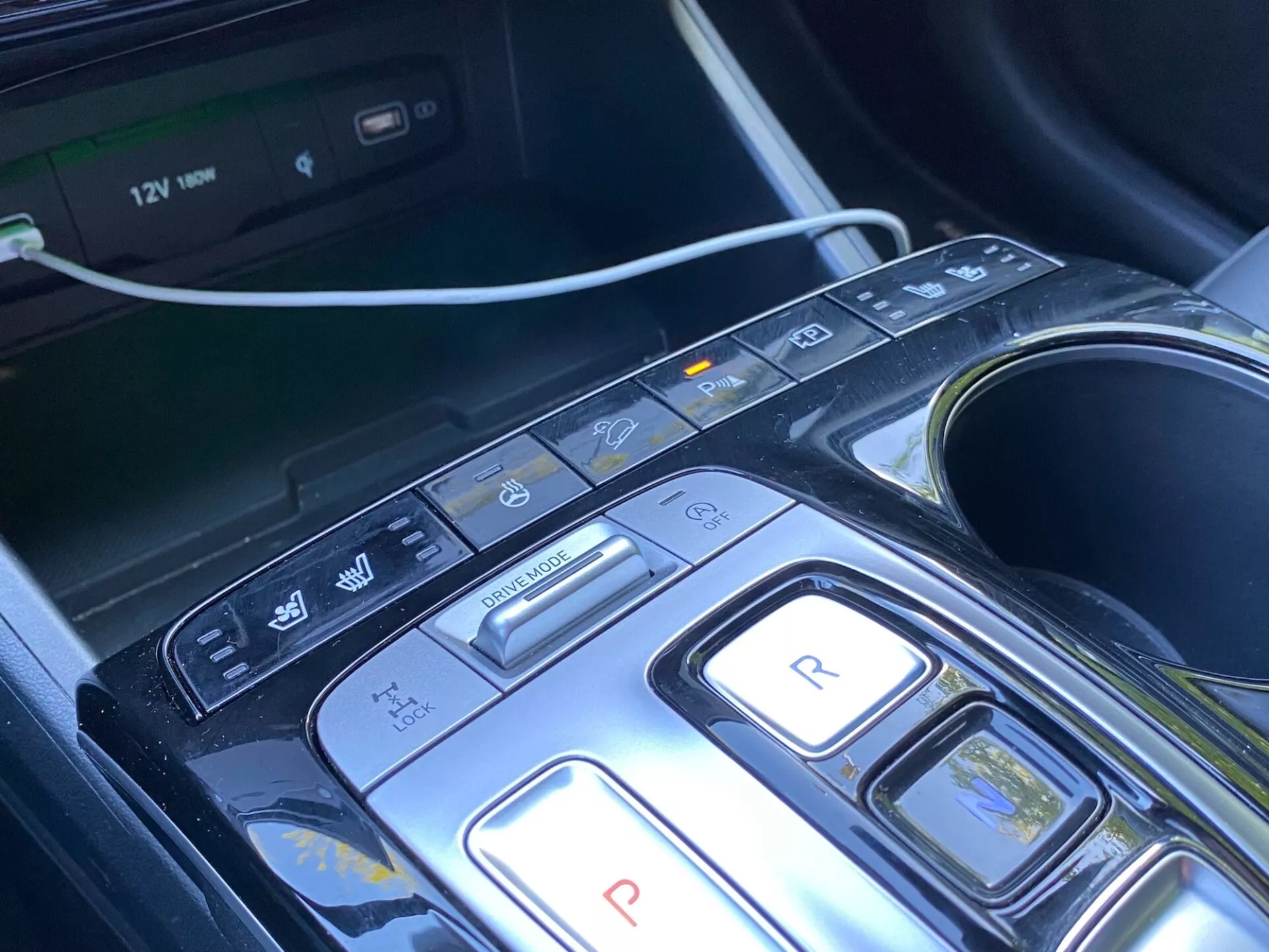 Primo piano della consolle centrale e dell'area di selezione del cambio di una Hyundai Tucson 2020 1.6 T-GDi Pure, con un design moderno con pulsanti per parcheggio, retromarcia, folle, guida e modalità freno/rigenerativa, insieme a pulsanti aggiuntivi per auto start/stop, sensori di parcheggio/parcheggio automatico e avviso di mantenimento/uscita di corsia. Display del sistema multimediale visibile, portabicchieri e presa di corrente 12V che indicano comfort, convenienza e sistemi avanzati di assistenza alla guida (ADAS). La console ha un design elegante con materiale nero lucido e accenti metallici, suggerendo un'appeal di lusso.