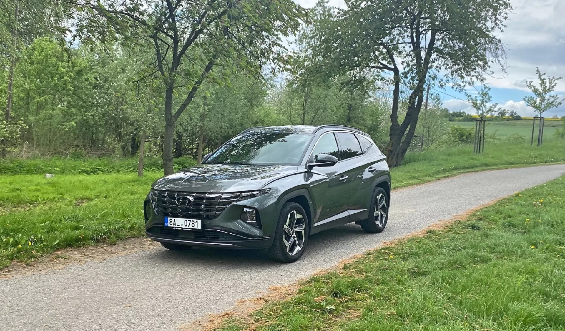 Vista anteriore di tre quarti di un crossover SUV Hyundai Tucson 2020 parcheggiato su un sentiero pavimentato con verde sullo sfondo, che mostra la distintiva griglia anteriore del veicolo, i moderni fari a LED, le grandi ruote in lega multiraggio, la linea del tetto inclinata, l'altezza da terra elevata, gli specchietti laterali con indicatori di direzione integrati, i vetri oscurati e i contorni stilistici del corpo.