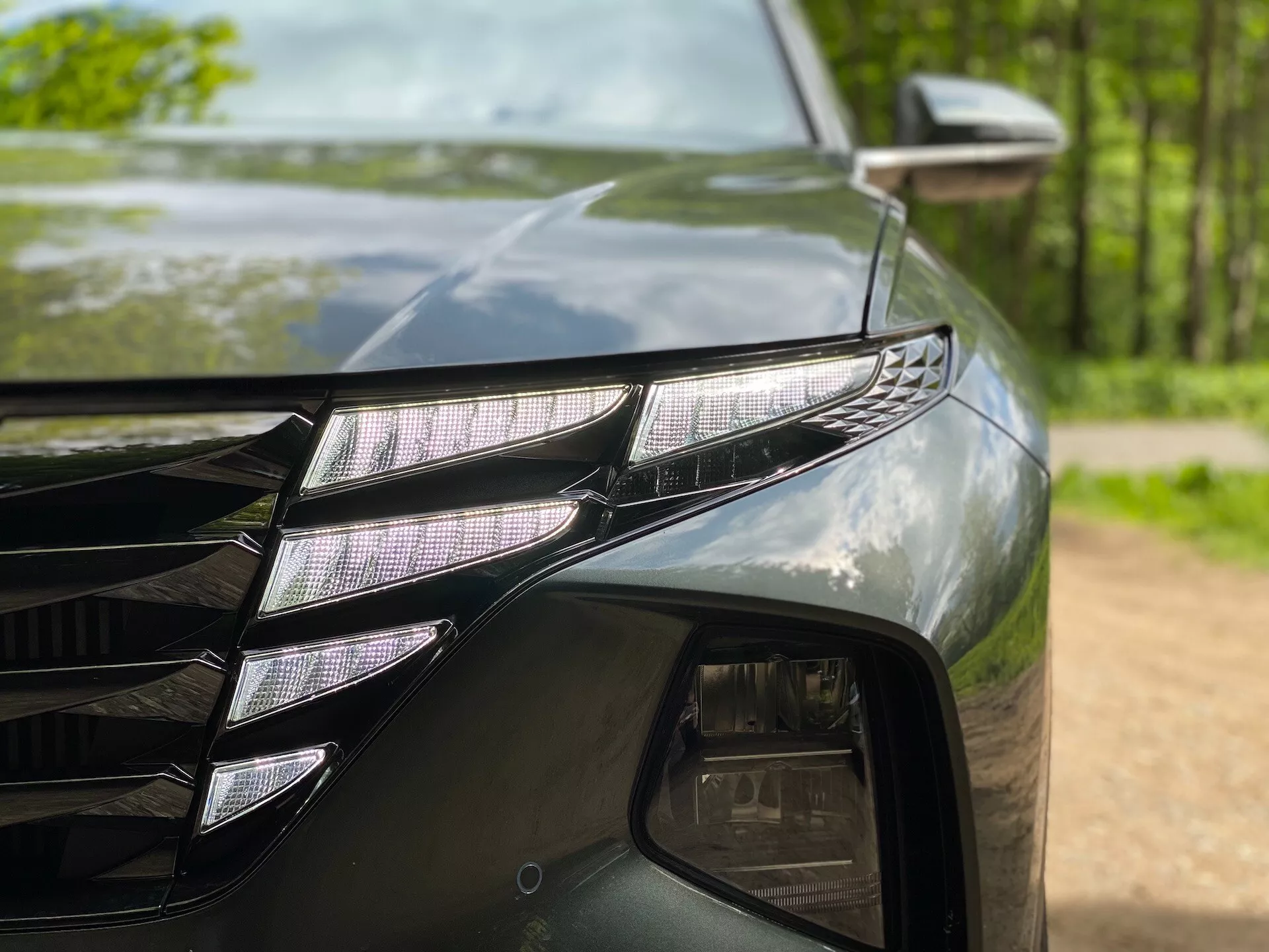 Primo piano del lato anteriore sinistro di una Hyundai Tucson 2020 (Hyundai-Tucson-1-6-T-GDi-Pure), che mostra un gruppo ottico LED con proiettori LED individuali e una distintiva striscia LED DRL. La vernice scura e lucida riflette gli alberi circostanti. È visibile anche una parte della griglia con linee angolari e lo specchietto laterale con indicatore di direzione.