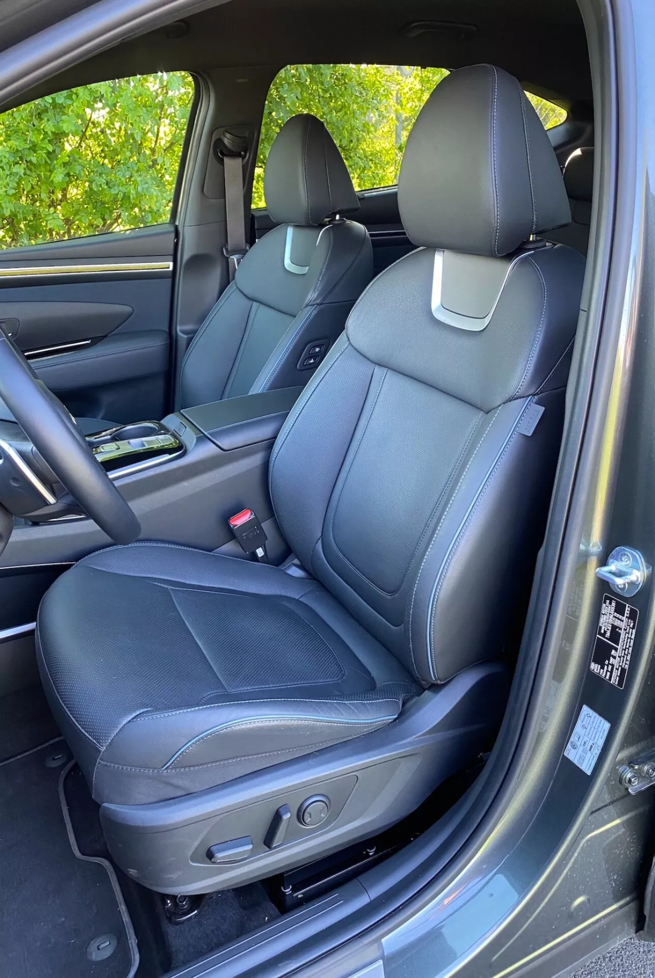 Interni del Hyundai Tucson 2020 1.6 T-GDI Pure che mostrano il sedile anteriore del passeggero con tappezzeria in pelle nera, cuciture dettagliate e comandi di regolazione elettrica. Consolle centrale visibile con portabicchieri, pannello della portiera con materiali coordinati, maniglia, vano portaoggetti e griglia dell'altoparlante che suggerisce un sistema audio avanzato. Cruscotto e vano portaoggetti parzialmente visibili. Tappetino di moquette e leggera riflessione della luce solare sul pannello della portiera.