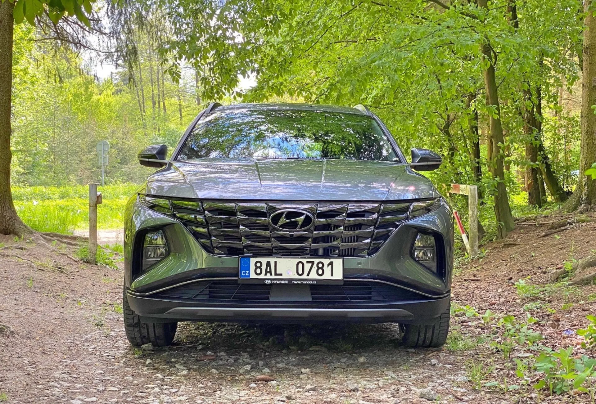 Vista frontale del veicolo Hyundai Tucson 1.6 T-GDi Pure 2020, che mostra una ripresa dal basso focalizzata sulla griglia, sul paraurti anteriore e sull'area del cofano. L'auto è parcheggiata su un sentiero ghiaioso in un ambiente naturale, suggerendo la sua idoneità per ambienti esterni. Le caratteristiche principali includono un design della griglia prominente, fari a LED eleganti e il logo Hyundai visibile.