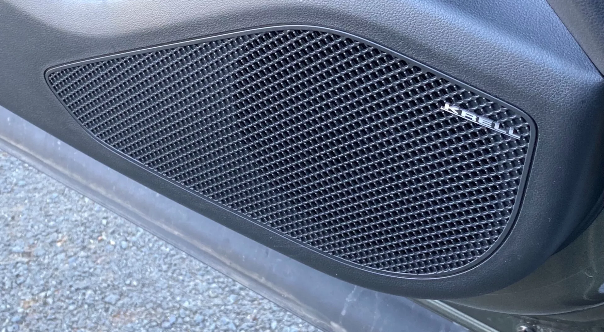 Vista laterale ravvicinata della Hyundai Tucson 2020 1.6 T-GDI Pure, concentrata su una griglia laterale con un motivo a rete con ritagli a forma di diamante. La griglia, incassata in un contorno sagomato, è modellata nel pannello della carrozzeria dell'auto con una finitura opaca, circondata da una superficie curva e liscia. L'immagine, catturata con un'angolazione obliqua dal basso verso l'alto, mostra un'illuminazione naturale all'aperto con pezzi di asfalto e detriti stradali, migliorando l'aspetto elegante e orientato alle prestazioni.