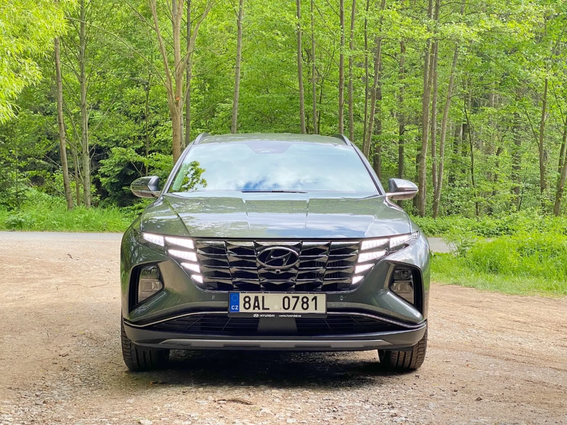 Vista frontale di un Hyundai Tucson 2020 (Hyundai Tucson 1.6 T-GDI Pure) SUV moderno parcheggiato su un sentiero ghiaioso con uno sfondo boscoso. L'immagine cattura lo stile elegante e aggressivo con una grande griglia geometrica che presenta il logo del produttore, fari LED angolari stretti e un paraurti anteriore scolpito con prese d'aria e recessi per i fari fendinebbia che evidenziano l'aerodinamica avanzata e la tecnologia di illuminazione.