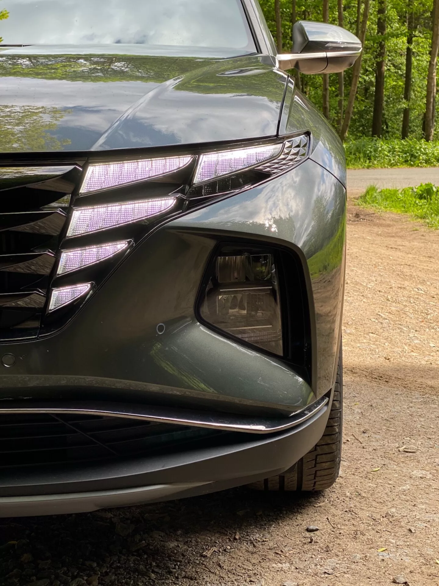Vista ravvicinata dell'angolo anteriore sinistro di una Hyundai Tucson 2020 1.6 T-GDI Pure, che mostra il faro con luci diurne a LED e lente stile proiettore, parte della griglia anteriore con linee orizzontali e accenti cromati, sezione di presa d'aria con alloggiamento per fendinebbia, paraurti anteriore scolpito con linee fluide e indicatore di direzione integrato nello specchio esterno. La vernice scura e lucida enfatizza le curve aerodinamiche della vettura.