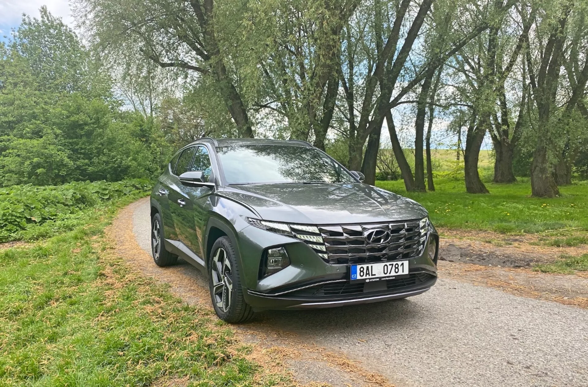 Vista frontale tre quarti di un Hyundai Tucson 1.6 T-GDi Pure 2020, che mostra caratteristiche moderne da SUV come fari a LED eleganti, griglia anteriore prominente con design geometrico, cofano aerodinamico scolpito, cerchi in lega, specchietti laterali con indicatori di direzione integrati e vernice metallica, sullo sfondo di una strada asfaltata stretta circondata da alberi, evidenziando la sua versatilità per ambienti urbani e rurali.