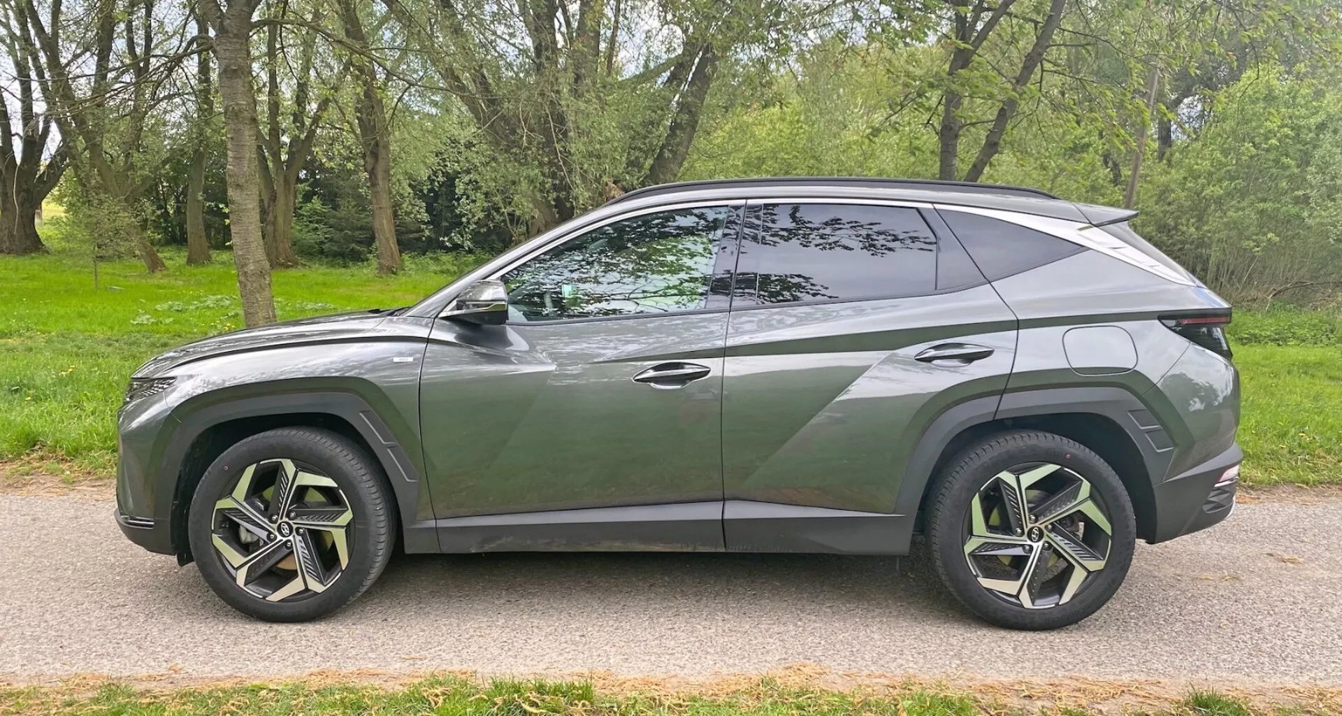 Profilo laterale del moderno SUV Hyundai Tucson 1.6 T-GDI Pure 2020 parcheggiato su una strada asfaltata accanto a un'area erbosa con alberi. Mostra un design aerodinamico ed elegante con una linea del tetto discendente, una linea del corpo distintiva, grandi cerchi in lega multirazze con pneumatici a basso profilo, finestrini oscurati con un montante B visibile, maniglie delle portiere a filo, specchietti laterali del colore della carrozzeria e finiture cromate lungo il bordo della finestra. Il veicolo cattura uno stile contemporaneo e possibili caratteristiche avanzate da un'angolazione laterale.