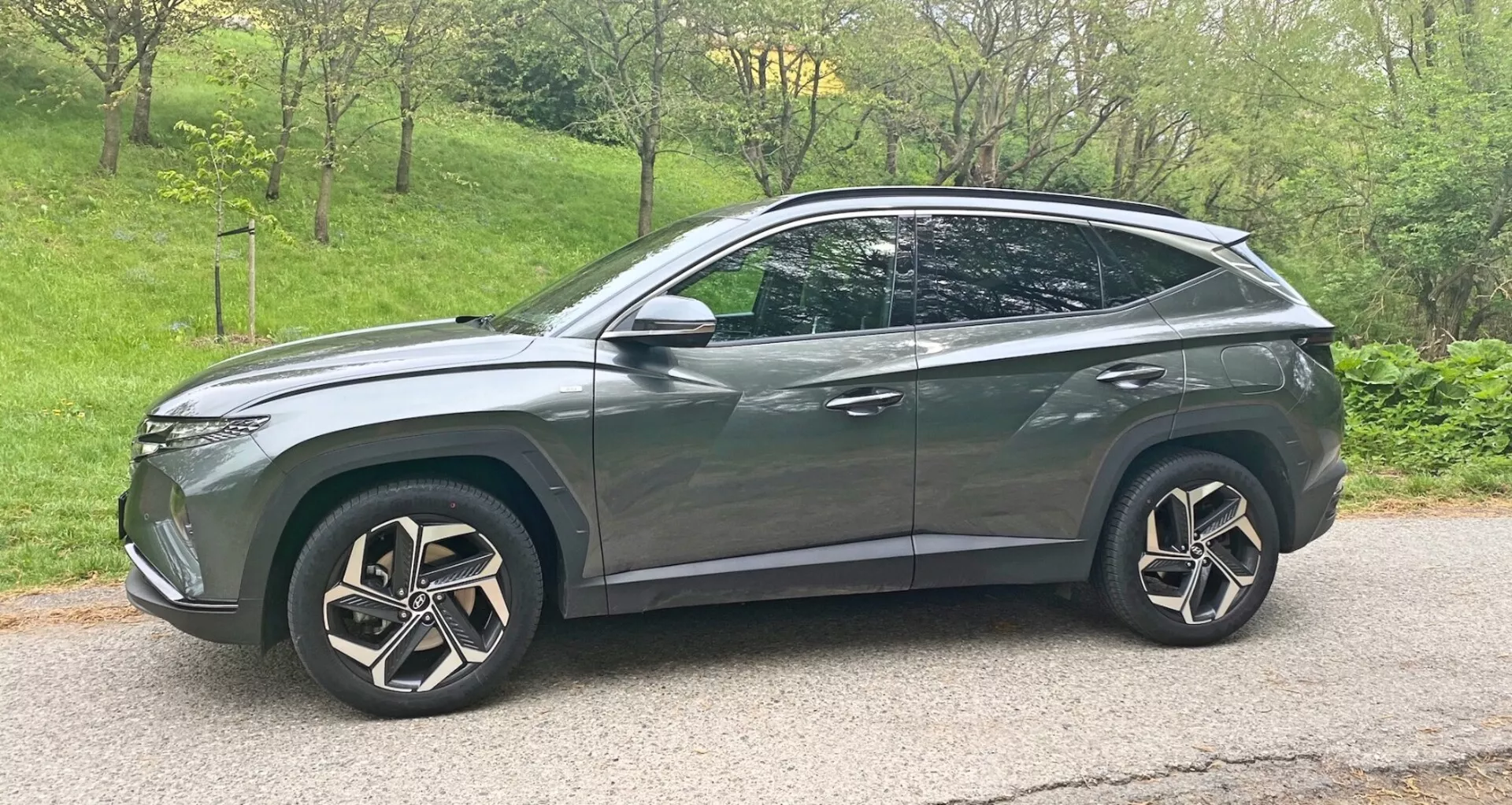 Profilo laterale di un moderno crossover SUV Hyundai Tucson 2020 1.6 T-GDI Pure in grigio metallico con finitura lucida. Il veicolo presenta grandi ruote in lega multirazze intricate, fari a LED, finestrini leggermente oscurati, linee aerodinamiche con tetto inclinato che termina in uno spoiler posteriore, barre sul tetto, specchietti laterali con indicatori di direzione integrati e accenti cromati o metallici. Il veicolo è posizionato su una strada asfaltata con un leggero angolo discendente e spostato verso destra dell'immagine.