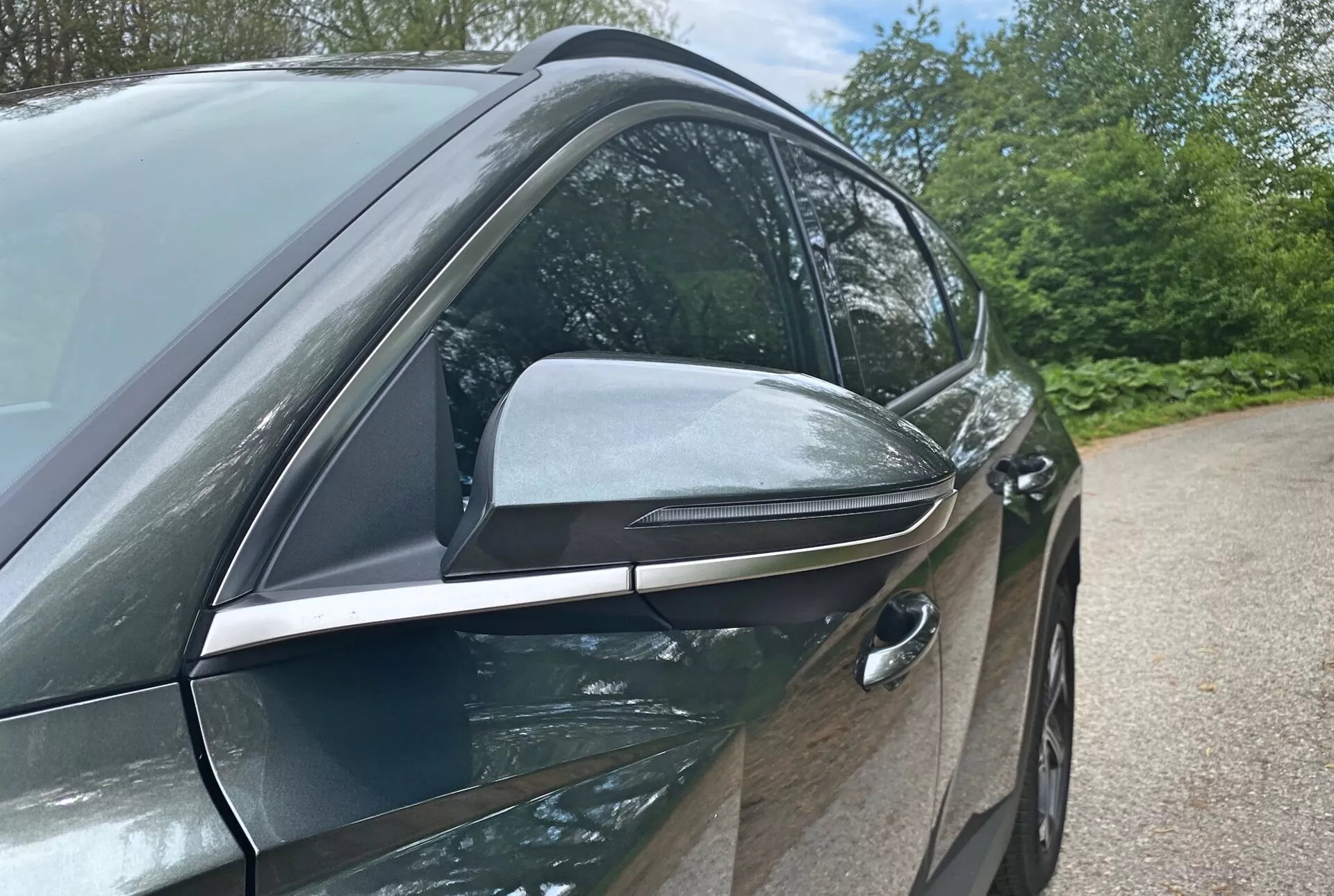 Vista laterale ravvicinata della Hyundai Tucson 2020 1.6 T-GDI Pure che mette in evidenza il design moderno e le caratteristiche tecnologiche, concentrandosi sul lato del guidatore. La fotografia a basso angolo sottolinea il design elegante e aerodinamico dello specchietto laterale con indicatori di direzione integrati e accenti neri, suggerendo funzionalità avanzate. La maniglia della portiera anteriore del guidatore a filo indica un sistema di accesso senza chiave, contribuendo all'efficienza aerodinamica del veicolo. Le porte di colore carrozzeria lucido e le finiture cromo delle finestre suggeriscono un pacchetto di finiture premium. Gli elementi di sfondo come alberi e cielo suggeriscono un ambiente esterno.