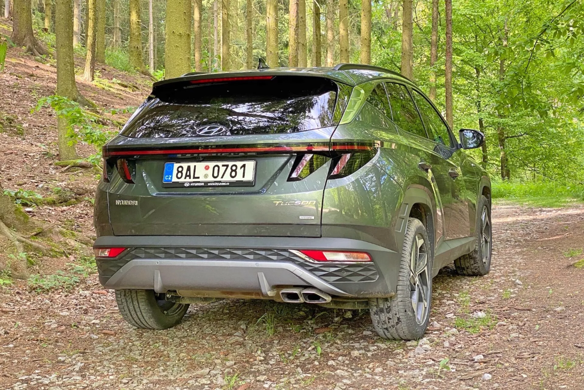 Immagine di un moderno SUV Hyundai Tucson 2020 in un ambiente boschivo con sentiero ghiaioso, catturata da un angolo posteriore di tre quarti. La foto mette in evidenza il lato destro e il profilo posteriore del veicolo con il portellone posteriore visibile, luci posteriori a LED, sistema di scarico doppio, spoiler posteriore e cerchi in lega.