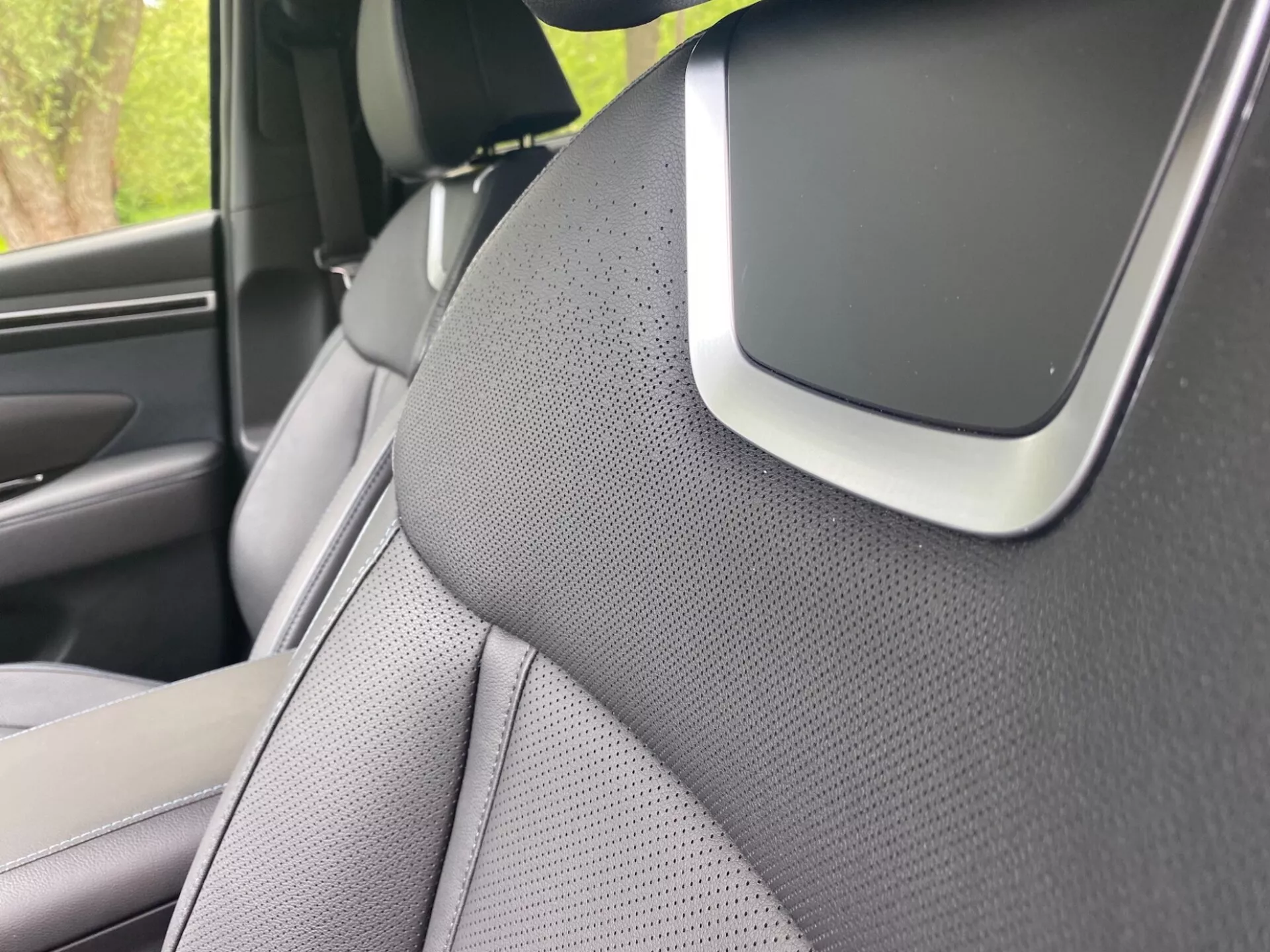 Primo piano dell'interno della Hyundai Tucson 2020, focalizzato sul montante A destro e parte del sedile anteriore destro. Il montante A è rivestito con un materiale testurizzato, probabilmente tessuto o pelle sintetica, suggerendo un'attenzione estetica o alla riduzione del rumore. Il sedile ha un design bicolore ed è perforato per la ventilazione. Una caratteristica notevole all'incrocio tra il montante A e il rivestimento del tetto potrebbe essere parte del sistema audio, un componente dell'airbag o un sensore di assistenza alla guida, evidenziando funzionalità avanzate di sicurezza o tecnologia.