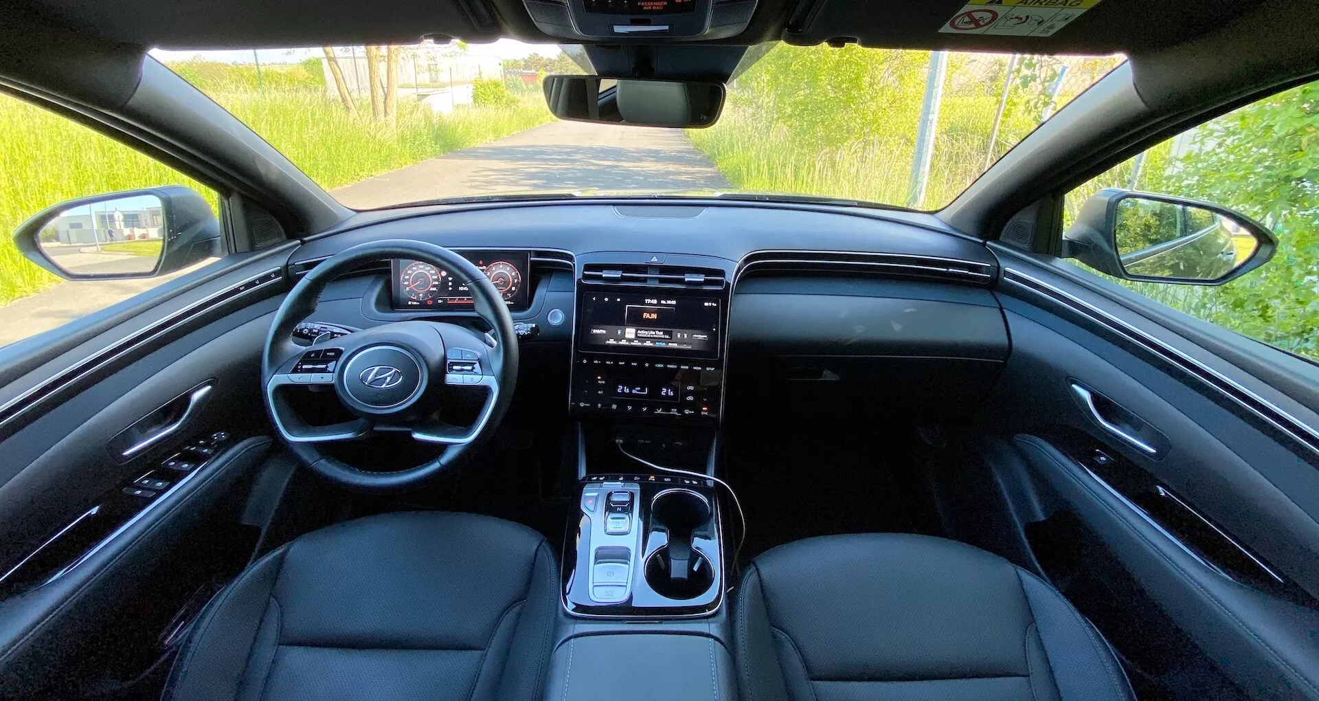 Vista dell'interno della Hyundai Tucson 2020 dal sedile posteriore che mostra il cruscotto anteriore, volante multifunzione con comandi integrati, quadro strumenti digitale, grande sistema di infotainment con schermo touch, pulsanti per il controllo del clima, leva del cambio e sedili anteriori in pelle sotto illuminazione naturale diurna.