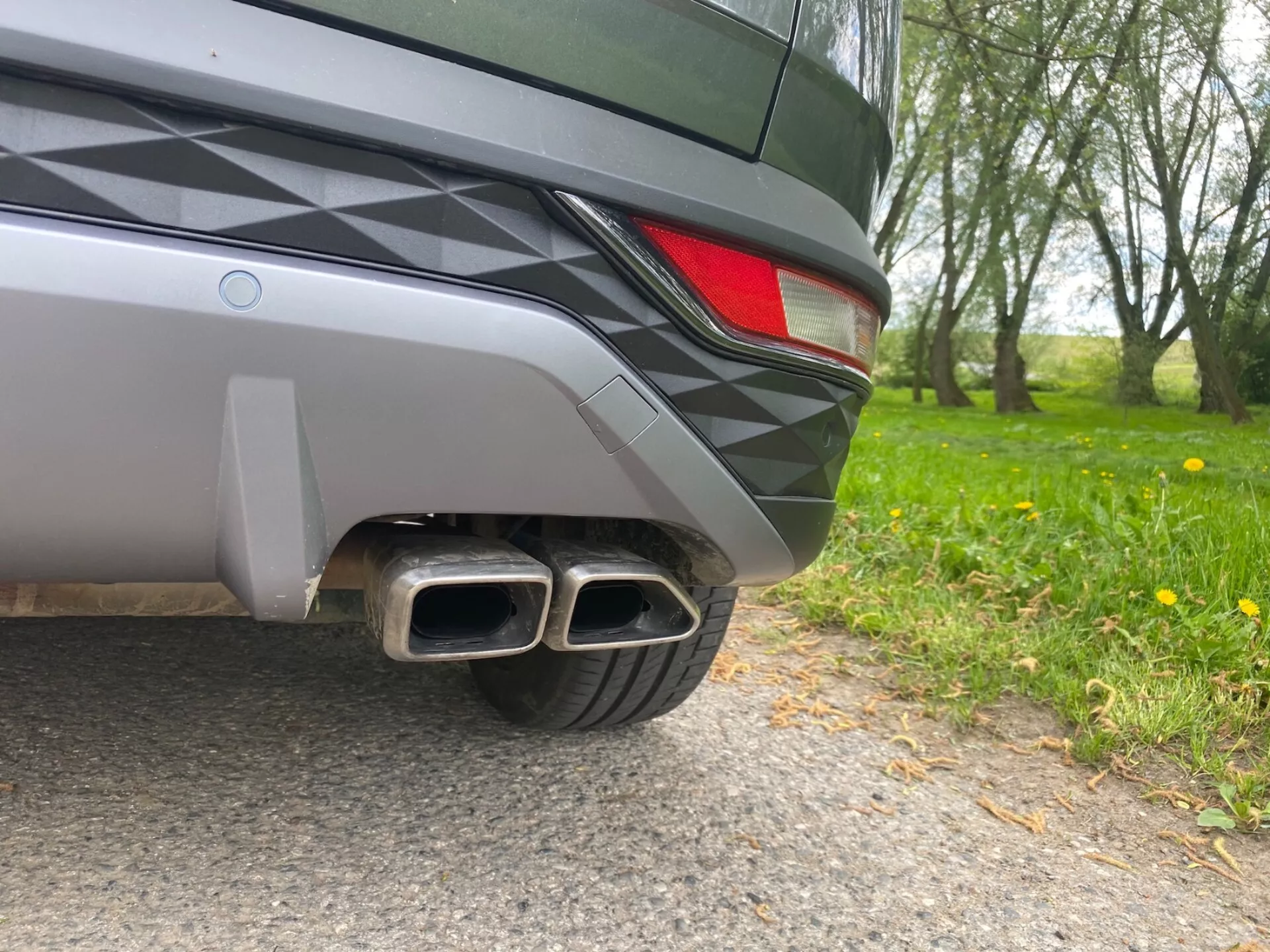 Immagine ravvicinata della sezione dell'angolo posteriore di una Hyundai Tucson 2020, con un angolo a livello del suolo. Dettagli visibili includono un paraurti posteriore bicolore, una parte del fanale posteriore a LED e doppi tubi di scarico metallici con punte rettangolari, suggerendo un allestimento sportivo. Il design elegante del veicolo e la vernice metallizzata riflettono l’ambiente naturale circostante.