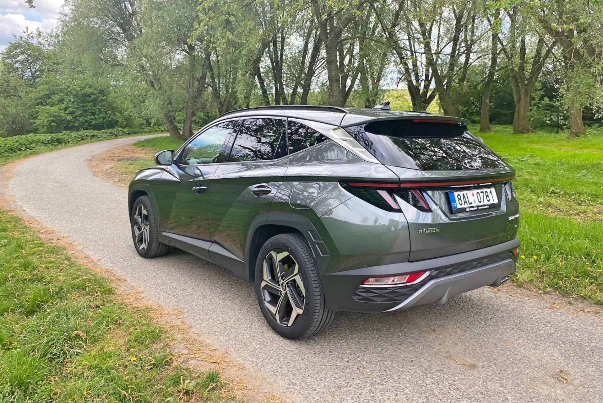 L'immagine mostra un SUV crossover moderno da un angolo posteriore di tre quarti, mettendo in evidenza il design aerodinamico e la linea del tetto inclinata. Il veicolo verde scuro presenta una porta posteriore in stile liftback, uno spoiler sportivo e grandi ruote in lega con design a più razze. Gli elementi visibili includono luci posteriori a LED moderne, doppi tubi di scarico e un tergicristallo posteriore. Il SUV è parcheggiato su un sentiero di ghiaia circondato dal verde, suggerendo un ambiente parco o rurale.