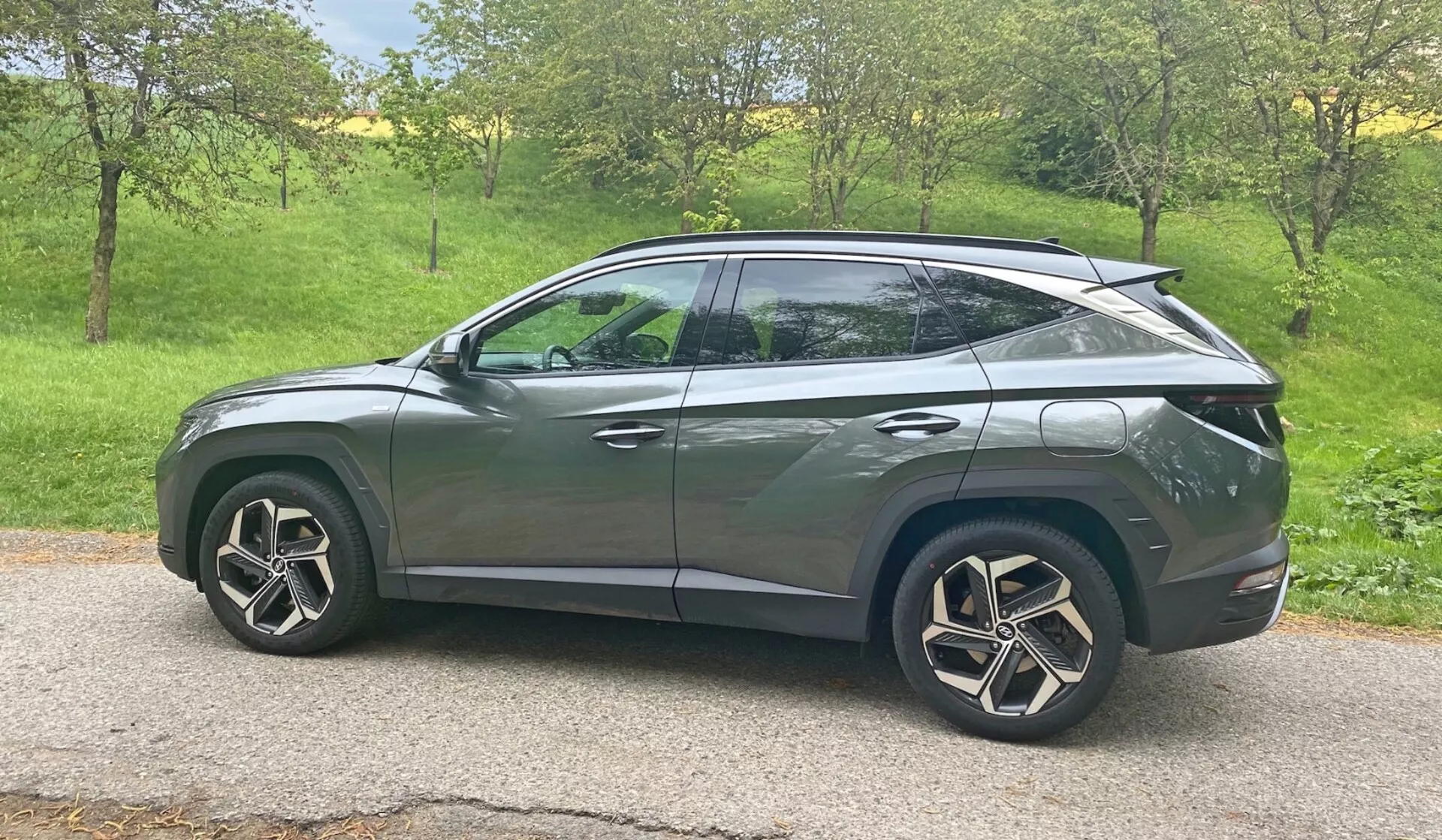 Vista laterale di un SUV Hyundai Tucson 1.6 T-GDI Pure del 2020 in un ambiente naturale alla luce del giorno. Il veicolo è posizionato in un'area erbosa con alberi sullo sfondo, suggerendo un'ambientazione rurale o suburbana. Le caratteristiche di design moderno includono fari LED, indicatori di direzione integrati negli specchietti laterali, vetri oscurati, cerchi in lega aerodinamici e una linea del tetto inclinata stile coupé. La carrozzeria elegante con linee caratteristiche e maniglie delle porte a filo suggeriscono un avanzato sistema di accesso senza chiave, mentre lo spoiler posteriore indica miglioramenti aerodinamici. L'auto è situata centralmente nell'inquadratura, offrendo una vista chiara e senza ostacoli da una prospettiva a metà altezza.