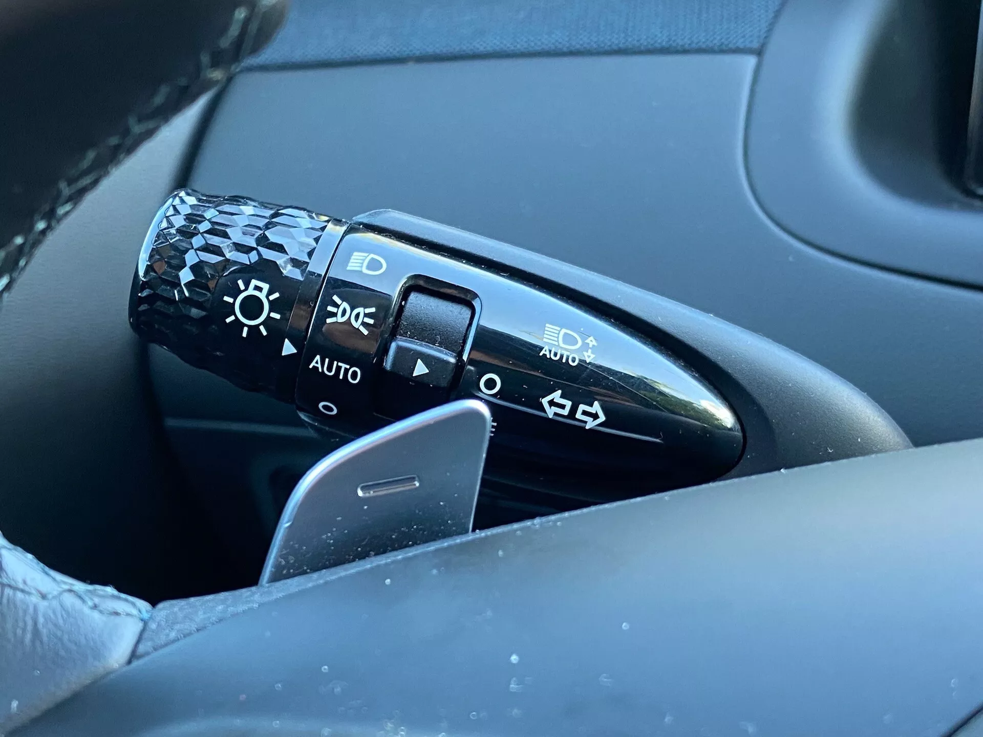 Vista ravvicinata della leva di controllo situata sulla colonna dello sterzo a sinistra del volante di una Hyundai Tucson 2020. La leva presenta simboli per le impostazioni dei fari tra cui spento, fari automatici, luci di parcheggio e fari abbaglianti/anabbaglianti. L'immagine cattura la leva, parte del volante e una porzione della porta lato guida in uno sfondo sfocato, concentrandosi sul dettaglio e la qualità del design. La posizione 'AUTO' indica un sistema di fari automatici.
