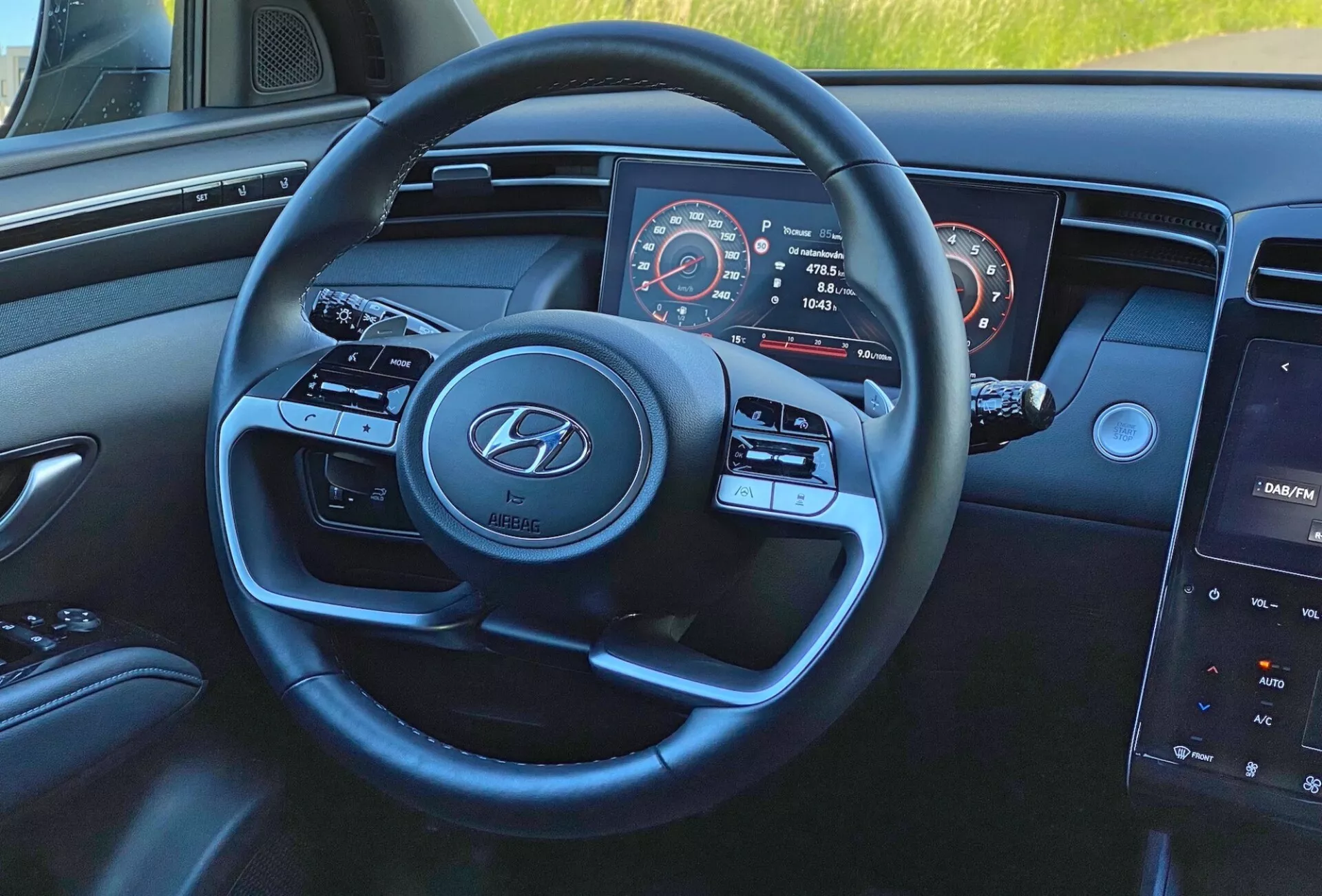 Vista interna del Hyundai Tucson 1.6 T-GDI 2020 dalla prospettiva del conducente, focalizzata sul volante a tre razze moderno con logo Hyundai, pulsanti per i media e il controllo della velocità, quadro strumenti parzialmente visibile con tachimetro e contagiri, e sistema di infotainment centrale con schermo touch sul cruscotto.