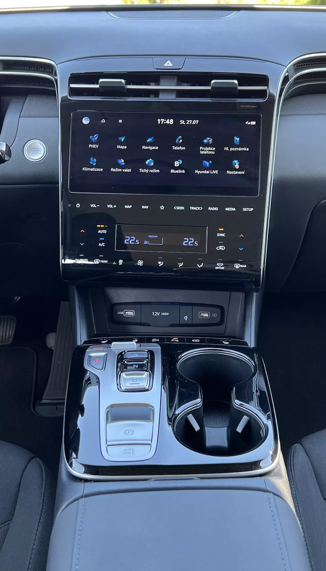 Interni della Hyundai Tucson 2020 che mostrano la console centrale con un grande sistema di infotainment touchscreen, pulsanti per il controllo del clima, selettore elettronico delle marce, selettore della modalità di guida, portabicchieri, vano portaoggetti e illuminazione ambientale.