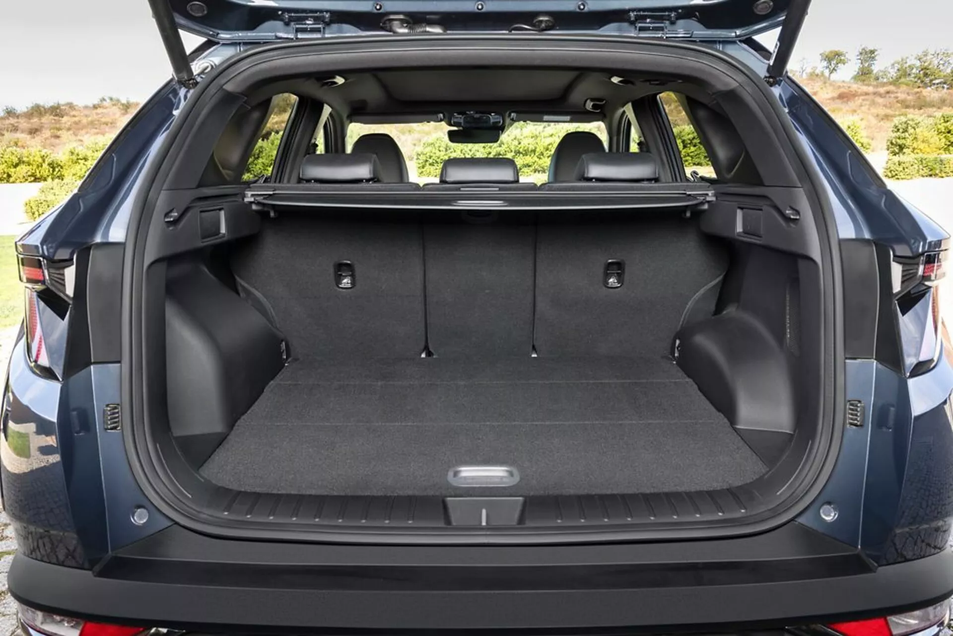 Area di carico posteriore di Hyundai Tucson 2020 con portellone aperto, che mostra pavimento di carico moquettato, punti di ancoraggio, vani di stoccaggio, presa da 12V, sedili posteriori ribaltabili, copertura del carico retrattile, pareti laterali in plastica resistente, e uno scorcio del paraurti posteriore, in un contesto esterno.