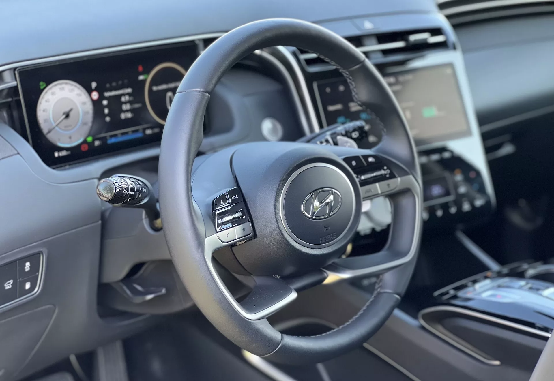 Vista interna della Hyundai Tucson 2020 focalizzata sul volante e sulle caratteristiche del cruscotto dalla prospettiva del conducente, evidenziando il design a tre razze con pulsanti di controllo, il quadro strumenti digitale e una vista parziale della console centrale e del controllo clima.