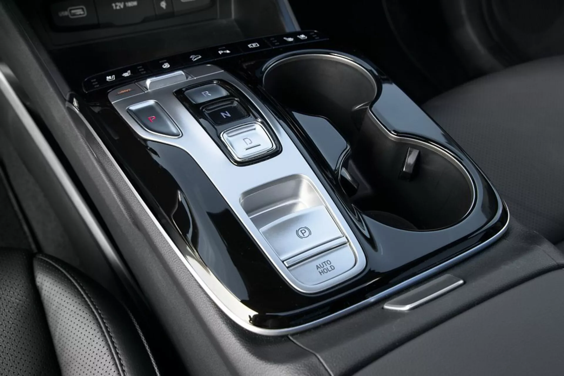 Primo piano della console centrale della Hyundai Tucson 2020 mostrando la leva del cambio automatico, pulsanti di controllo, funzione Auto Start/Stop, interruttore delle luci di emergenza e portabicchieri doppio con dettagli cromati, da una visuale dall'alto con leggera inclinazione.