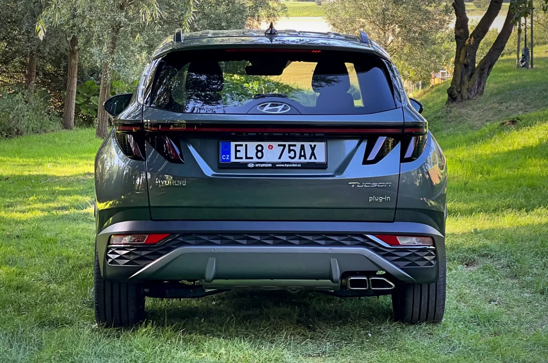 Vista posteriore della SUV Hyundai Tucson 2020 su pendio erboso con sfondo boscoso, mostrando logo sul portellone, badge Tucson e Hybrid, segno Plug-in, luci riflettenti, luci posteriori dinamiche, tergicristallo posteriore, luce di stop alta, punte di scarico doppie, piastre di scorrimento argento e badge AWD.