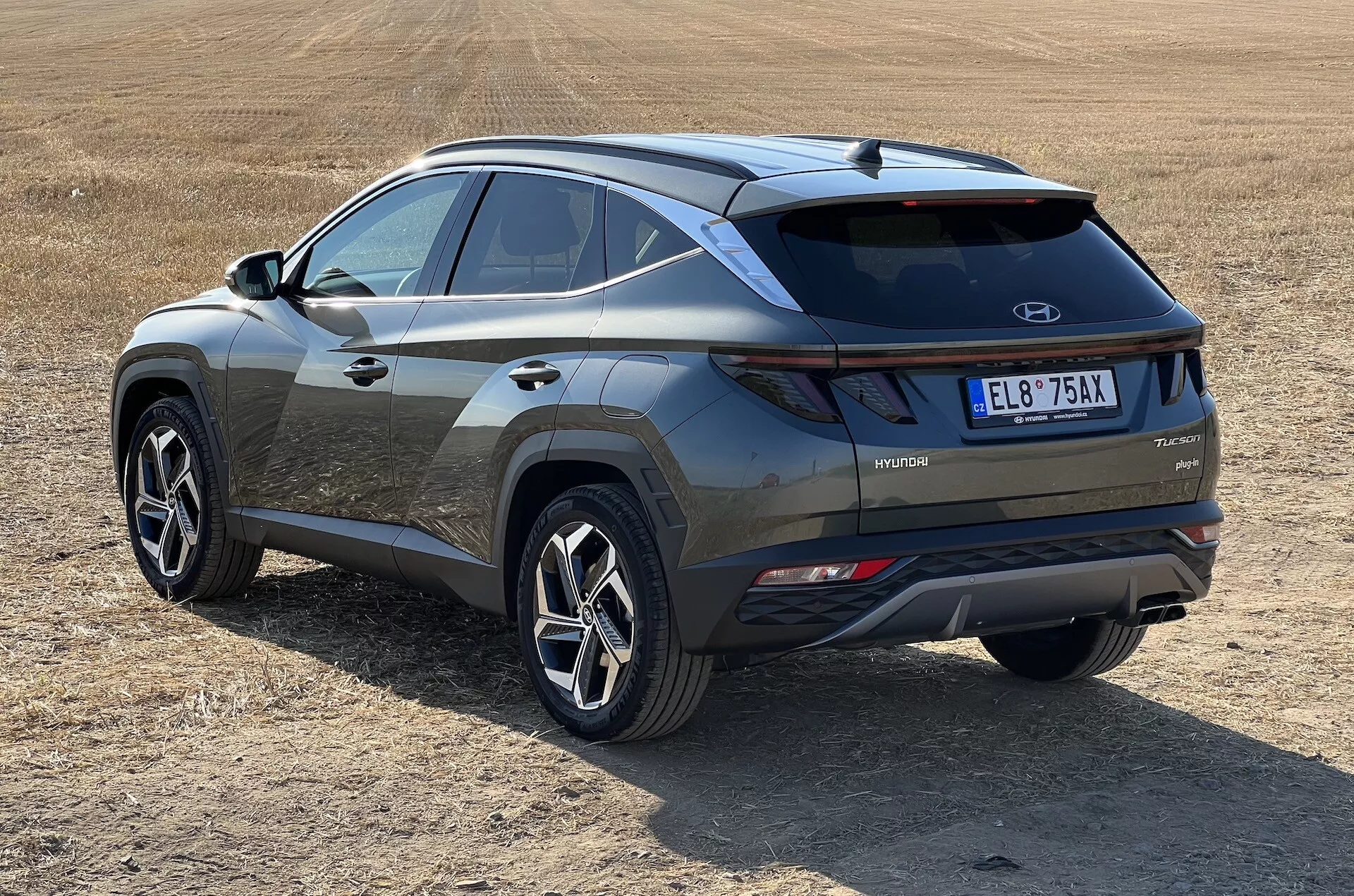 SUV moderno Hyundai Tucson 2020 in vista obliqua che mostra i profili posteriore e laterale con badge di tecnologia ibrida su campo erboso asciutto, evidenziando i fari posteriori LED, inserto paraurti a contrasto e ruote sportive a più razze