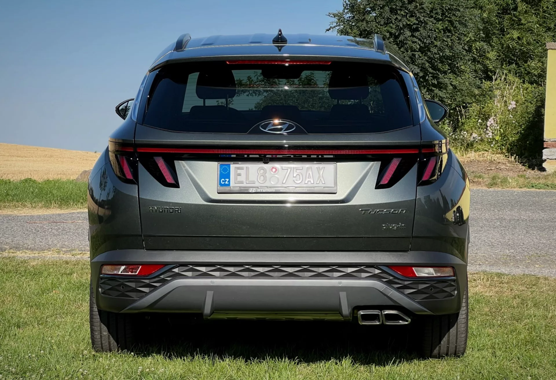 Vista posteriore di Hyundai Tucson 2020 che mostra fanali orizzontali, badge centrale, nome del livello di allestimento, sensori di parcheggio integrati nel paraurti e un sistema di scarico doppio, parcheggiata su una superficie fiancheggiata da erba contro un cielo sereno
