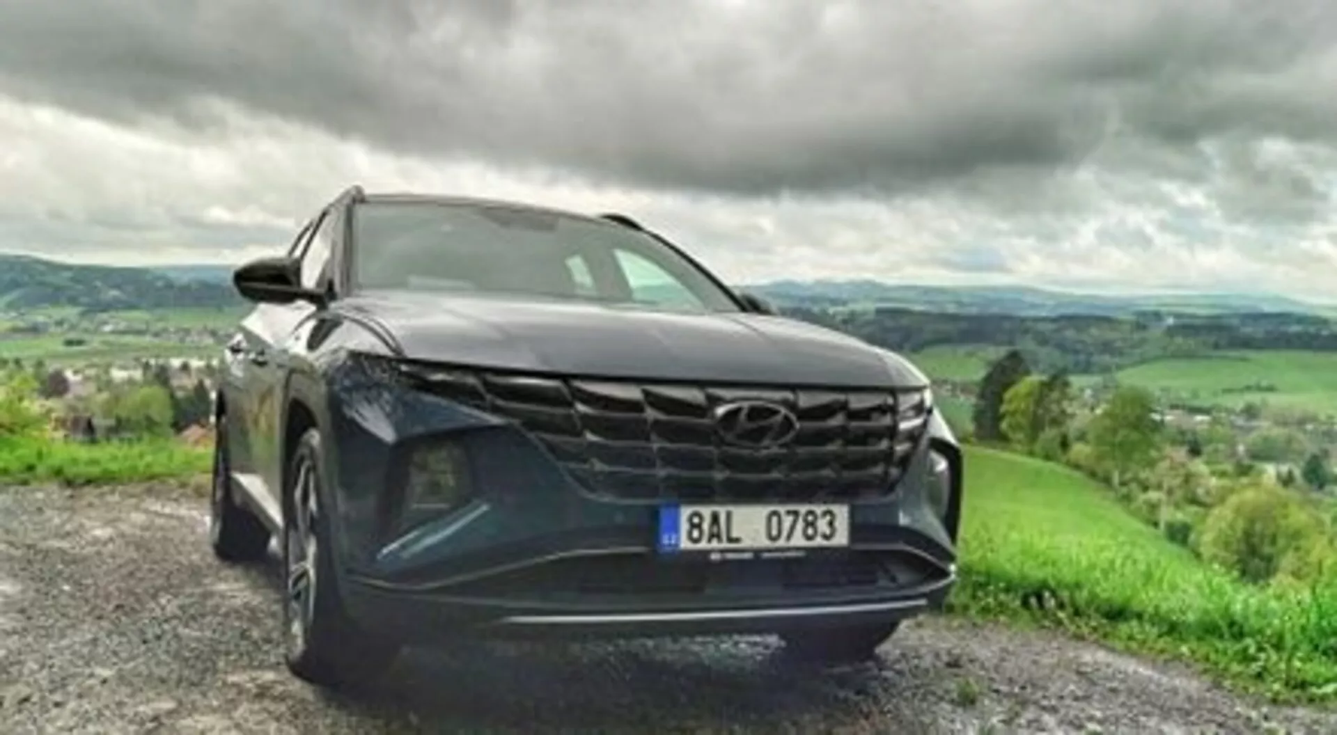 SUV Hyundai Tucson 2020 parcheggiata su strada sterrata, mette in mostra la sua griglia audace, fari LED eleganti e vernice metallica in una vista dinamica da un basso angolo frontale, con un paesaggio collinare lussureggiante sotto un cielo nuvoloso sullo sfondo