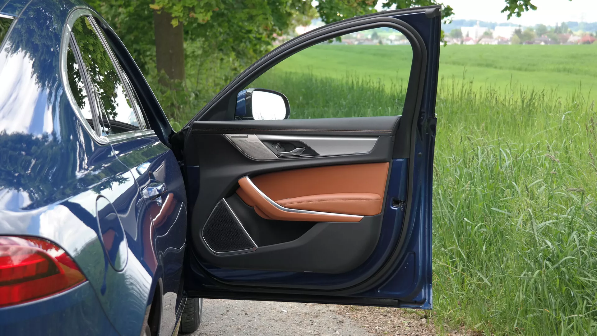 Vista laterale da vicino della Jaguar XF 2021 con la porta del conducente aperta, che rivela un interno lussuoso con una combinazione di pelle nera e marrone caldo. Lo specchio laterale, integrato con una freccia, corrisponde all'elegante esterno blu dell'auto. Parcheggiata su una superficie asfaltata vicino a un campo erboso, il parafango visibile parzialmente e il pannello posteriore evidenziano il design moderno e le caratteristiche tecnologiche avanzate.