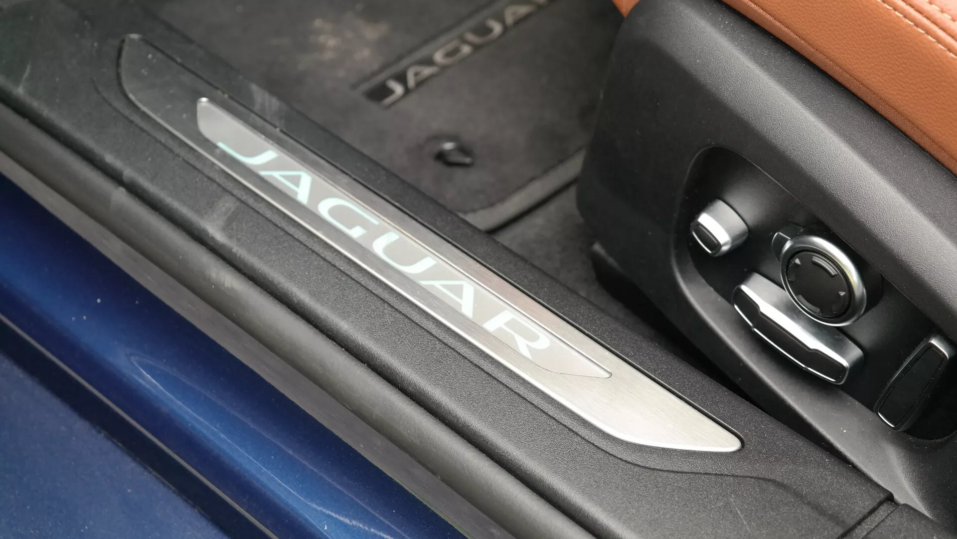 Dettaglio ravvicinato dell'interno della Jaguar XF 2021, con il marchio illuminato 'JAGUAR' sulla soglia della portiera in metallo spazzolato, accanto a un disegno a motivi circolari e un pulsante di controllo. Contrasti di texture tra finitura cromata, esterno blu visibile e sezione superiore in pelle in un ambiente veicolare lussuoso e high-tech.