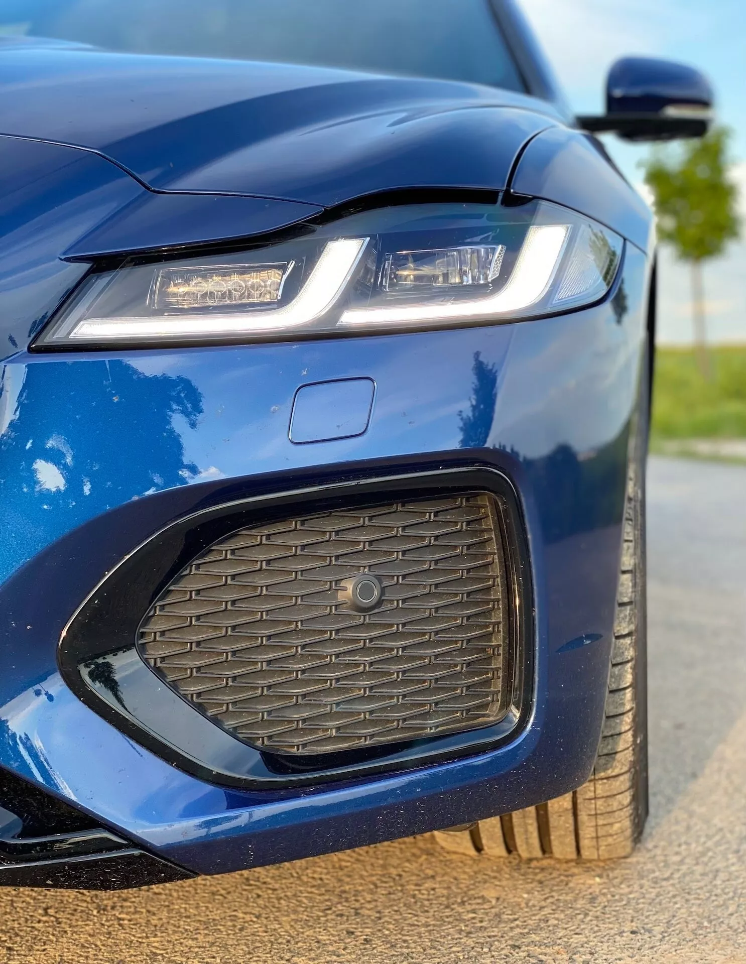 Immagine in primo piano della sezione anteriore sinistra della Jaguar XF 2021 in blu metallizzato. La ripresa cattura il faro sinistro con tecnologia di illuminazione LED o laser, una parte del cofano, il paraurti anteriore con una presa d'aria a nido d'ape, una sezione della griglia con un grande emblema e una parte dello specchietto laterale sinistro. L'immagine evidenzia il design affilato e aggressivo dell'auto, l'estetica sportiva, la finitura lucida, il design aerodinamico dello specchietto laterale con indicatori di direzione integrati e la ruota in lega parzialmente visibile, mostrando le caratteristiche di lusso e tecnologiche dell'auto.