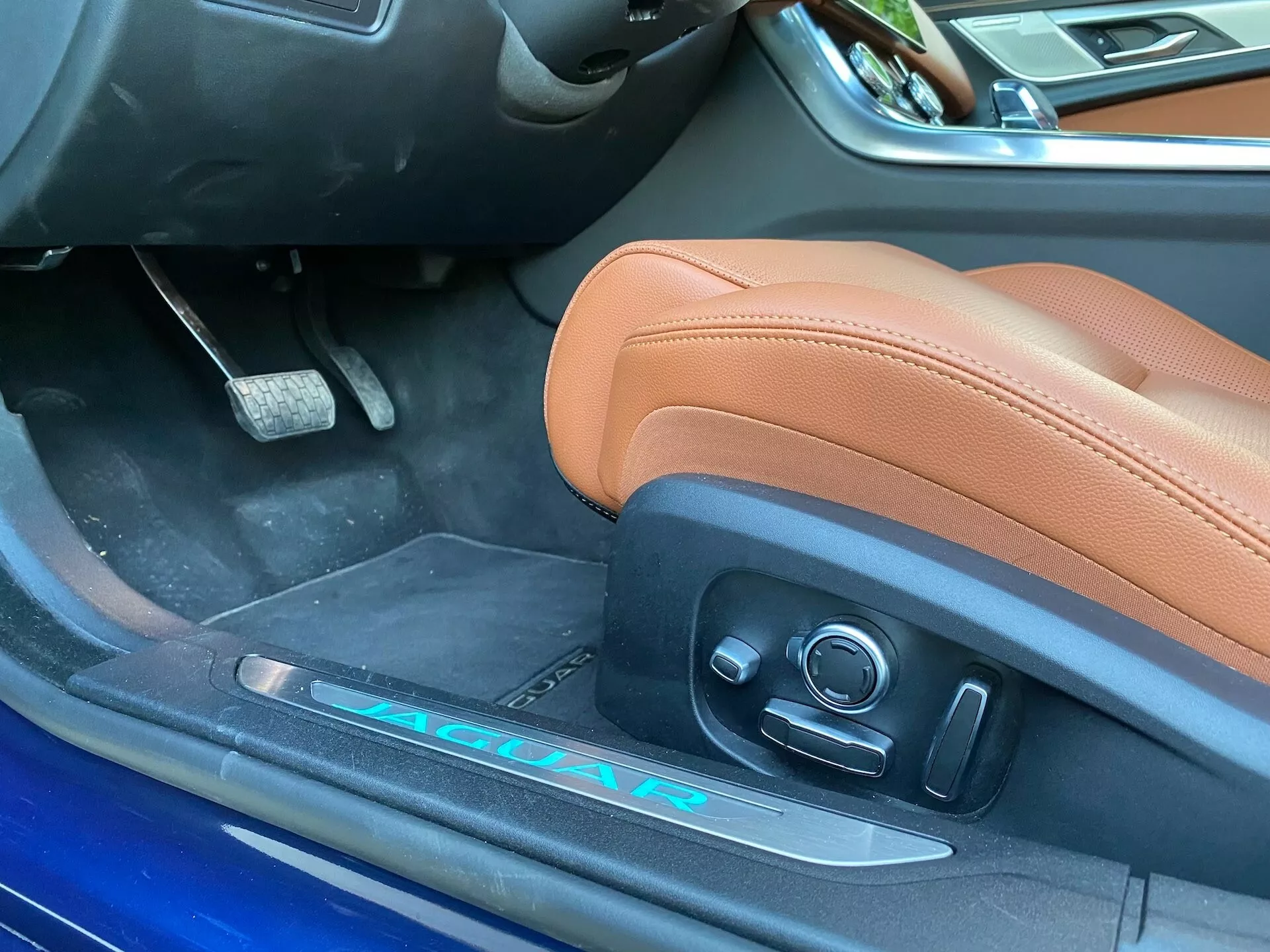 Vista ravvicinata dell'interno lato guidatore di una Jaguar XF 2021, che mostra un sedile in pelle marrone chiaro con comandi elettrici, battitacco in alluminio parzialmente visibile con marchio 'JAGUAR' e 'R' e bordo della porta blu vibrante. L'immagine cattura le caratteristiche lussuose e sportive del veicolo da un'angolazione leggermente inclinata vicino alla porta aperta.