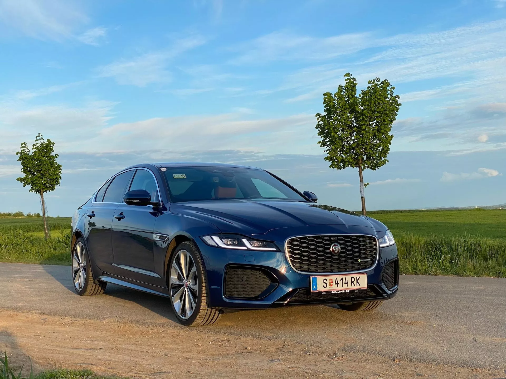 L'immagine mostra una berlina di lusso blu scuro, Jaguar XF 2021, da un angolo frontale basso di tre quarti. Posizionato su uno spiazzo di terra o ghiaia accanto a una strada asfaltata con prato e un cielo limpido e blu sullo sfondo, l'auto sfoggia i suoi fari a LED e la grande griglia distintiva anteriore con un badge centrale che indica il marchio. La prospettiva di tre quarti cattura il cofano, il parabrezza, la linea del tetto e il lato anteriore dell'auto, mostrando cerchi in lega con un design a più razze. Il cofano con linee scolpite, i vetri oscurati per la privacy e gli specchietti laterali con indicatori di direzione integrati suggeriscono un design dinamico e di lusso. L'illuminazione naturale, insieme a caratteristiche tecnologiche avanzate visibili come i sistemi di assistenza alla guida e le opzioni di infotainment, sottolineano il suo status di lusso.