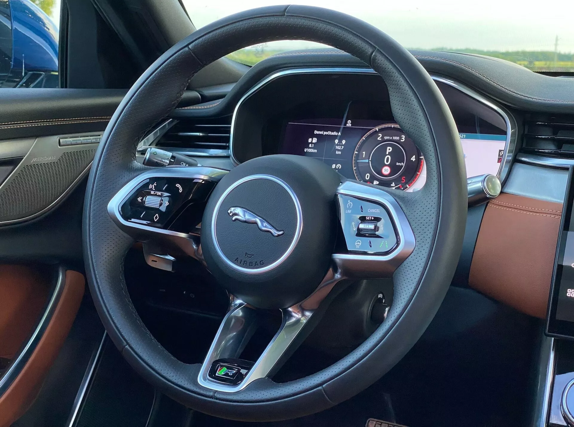 Vista interna della Jaguar XF 2021 dal posto di guida, con il volante a tre razze rivestito in pelle nera con cuciture visibili e il caratteristico logo del felino saltante. Il raggio sinistro contiene comandi per l'audio e i comandi vocali, mentre il raggio destro include pulsanti per il controllo della velocità e il sistema di informazioni del conducente. Il cruscotto dietro il volante mostra un mix di quadranti digitali e analogici, compreso il tachimetro e il contagiri. L'interno lussuoso mette in risalto il rivestimento in pelle bicolore in ricco marrone e nero.