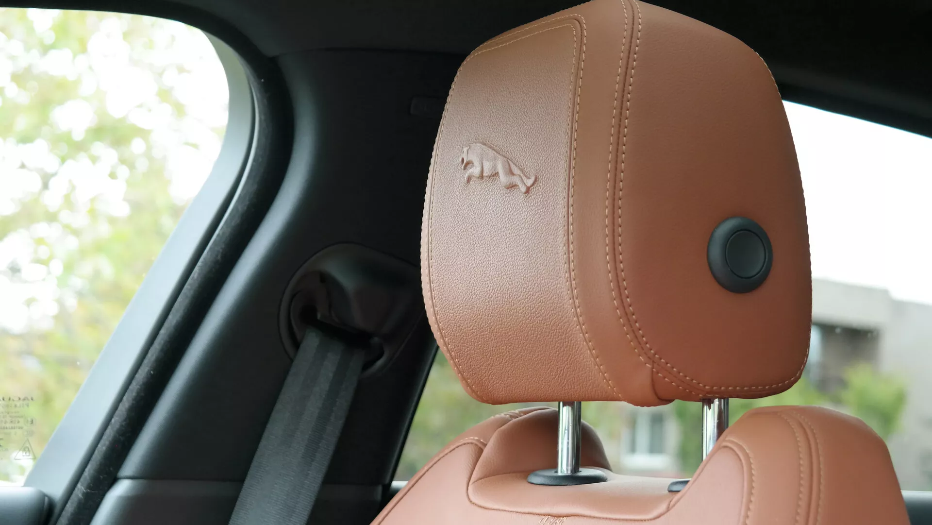 Vista interna della Jaguar XF 2021 con messa a fuoco sul poggiatesta lato guida, ripresa dalla prospettiva del sedile posteriore. Il poggiatesta, rivestito in pelle di alta qualità color marrone chiaro con cuciture visibili, presenta un logo in rilievo, un guida-cintura ergonomica e un meccanismo di regolazione manuale. Lo sfondo include un montante B sfocato e un finestrino, mettendo in evidenza il design lussuoso ed ergonomico dell'auto.