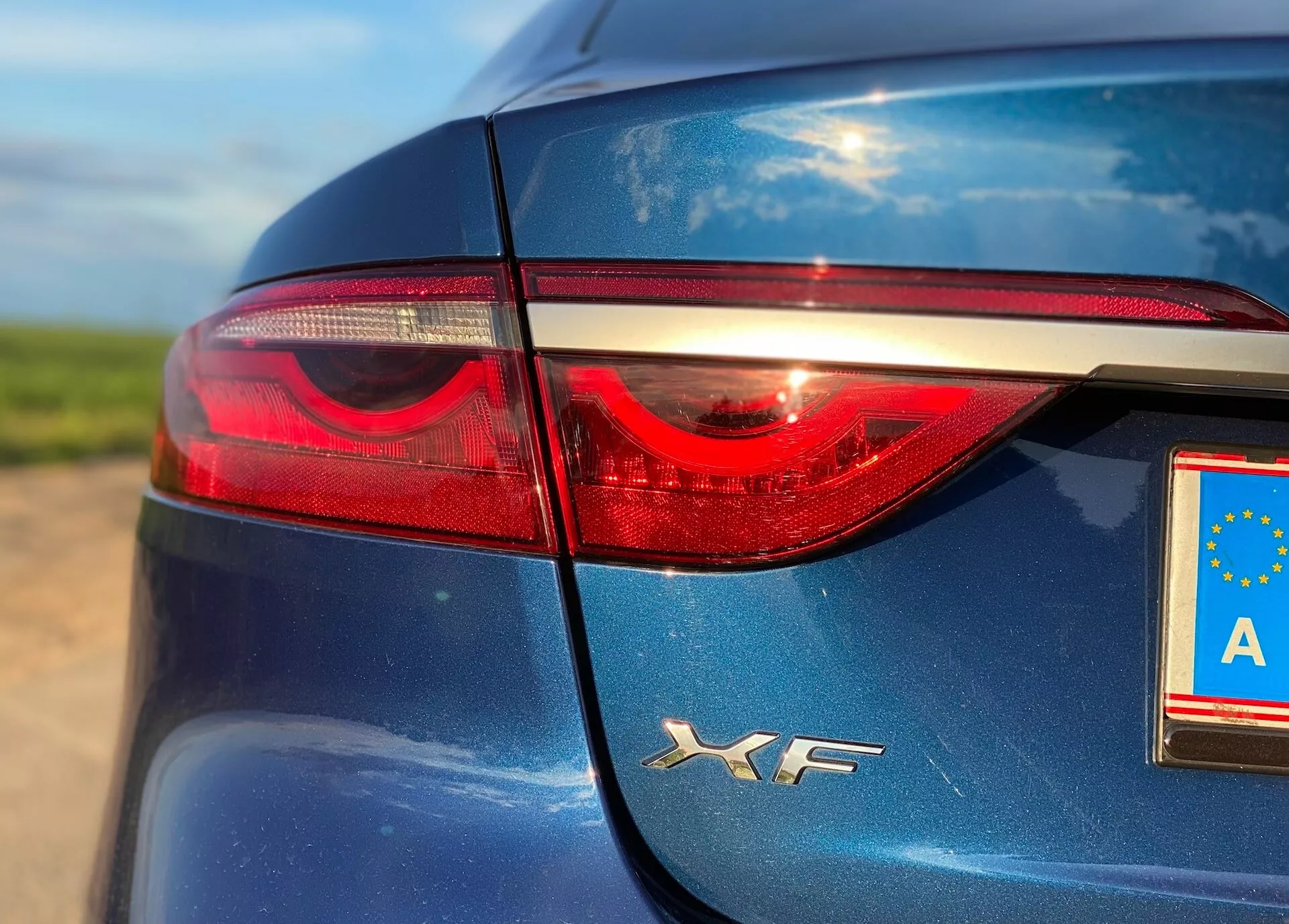 Immagine ravvicinata che mostra la sezione posteriore sinistra di una Jaguar XF 2021. La foto mette in evidenza il corpo elegante di colore blu scuro, la finitura lucida e la luce posteriore sinistra con una striscia di LED. La targa con il simbolo dell'UE presenta la lettera 'A' per l'Austria, con riflessi che indicano luce naturale.