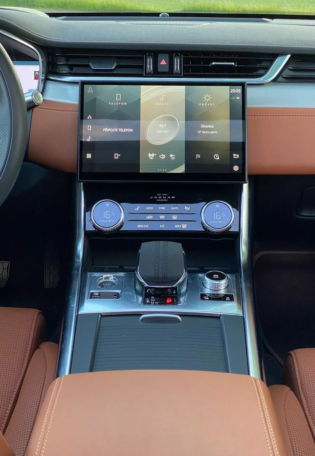 Interno della Jaguar XF 2021, mostrando una prospettiva centrata sul conducente dal sedile anteriore con attenzione al cruscotto e alle caratteristiche tecnologiche all'avanguardia. Spiccano un grande sistema di infotainment touchscreen user-friendly, un elegante pannello di controllo della climatizzazione bi-zona, un selettore del cambio elettronico, sedili in pelle di lusso con cuciture a contrasto e illuminazione ambientale. Sono visibili anche elementi pratici come i portabicchieri e i compartimenti di stoccaggio. La luce naturale che invade l'abitacolo suggerisce un tetto panoramico o un grande parabrezza, migliorando l'atmosfera lussuosa complessiva.