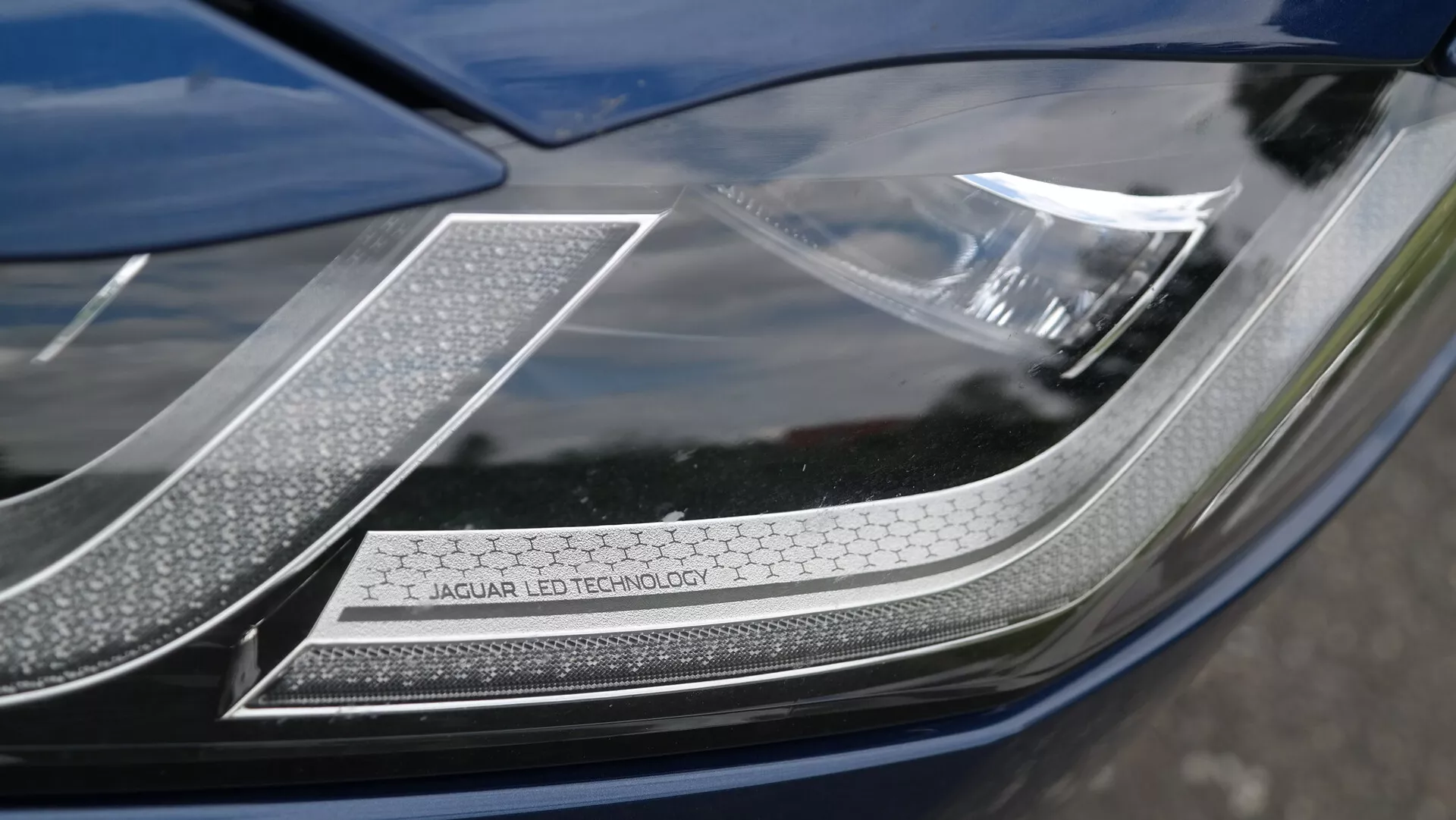 Primo piano del faro della Jaguar XF 2021, focalizzato sull'angolo in alto a destra da una prospettiva leggermente angolata. Caratteristiche includono l'iscrizione visibile 'JAGUAR LED TECHNOLOGY' su una piastra con finitura cromata, un intricato disegno dei LED e luci diurne con motivo a nido d'ape. Il faro presenta una copertura liscia e lucida con curvatura aerodinamica, evidenziando la tecnologia LED avanzata e il design premium di Jaguar.