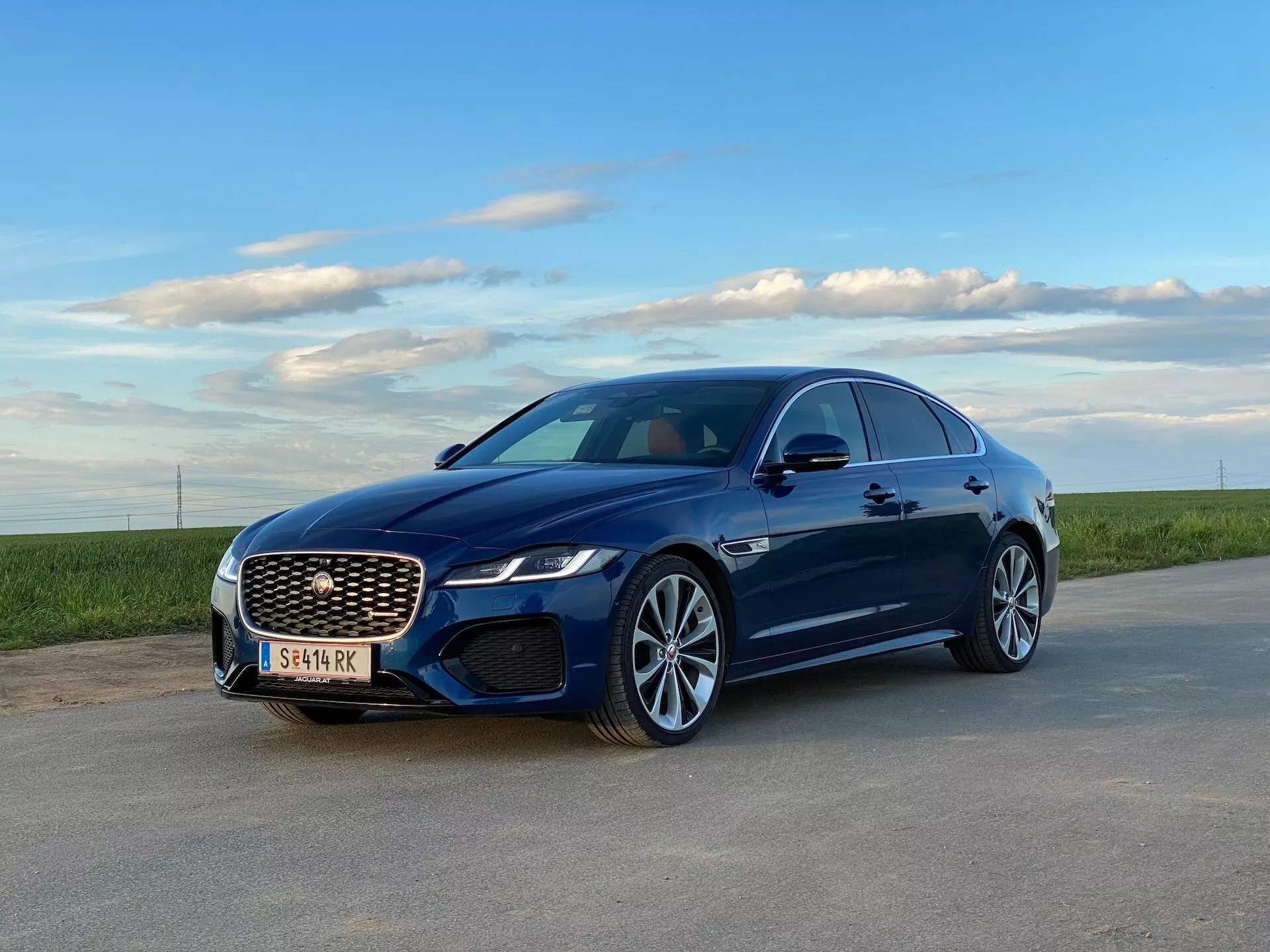 Una berlina di lusso blu fotografata da un angolo frontale di tre quarti, rivelando il suo elegante frontale e il profilo laterale. L'auto presenta linee di carrozzeria morbide, una grande griglia anteriore a rete, fari a LED e cerchi in lega multirazze. La prospettiva è leggermente verso l'alto, enfatizzando i dettagli della parte inferiore. L'immagine sembra essere stata scattata in una chiara serata presto su una strada asfaltata con un orizzonte erboso sullo sfondo. Si presume la presenza di vetri oscurati e una possibile gamma di caratteristiche tecnologiche avanzate come sensori di parcheggio e ADAS.