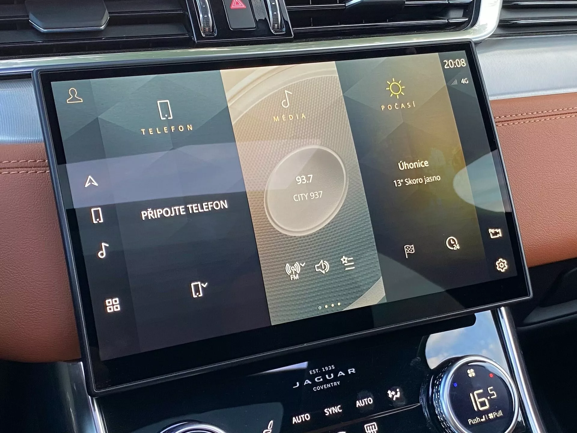Sistema di infotainment touchscreen della Jaguar XF 2021 posizionato nella console centrale, visto dalla prospettiva di un passeggero o guidatore. L'attenzione è sullo schermo, che mostra più icone per la connettività telefonica, la riproduzione multimediale e le applicazioni specifiche del veicolo, tra cui Radio, Media e Podcast, con segni di integrazione con lo smartphone. Interfaccia moderna touch-based dal design elegante, con impostazioni di sistema visibili come l'ora (20:08) e AUTO per il controllo del clima. Sotto, sono visibili una manopola rotante e i controlli del clima a doppia zona con una regolazione della temperatura di 16,5 gradi Celsius.