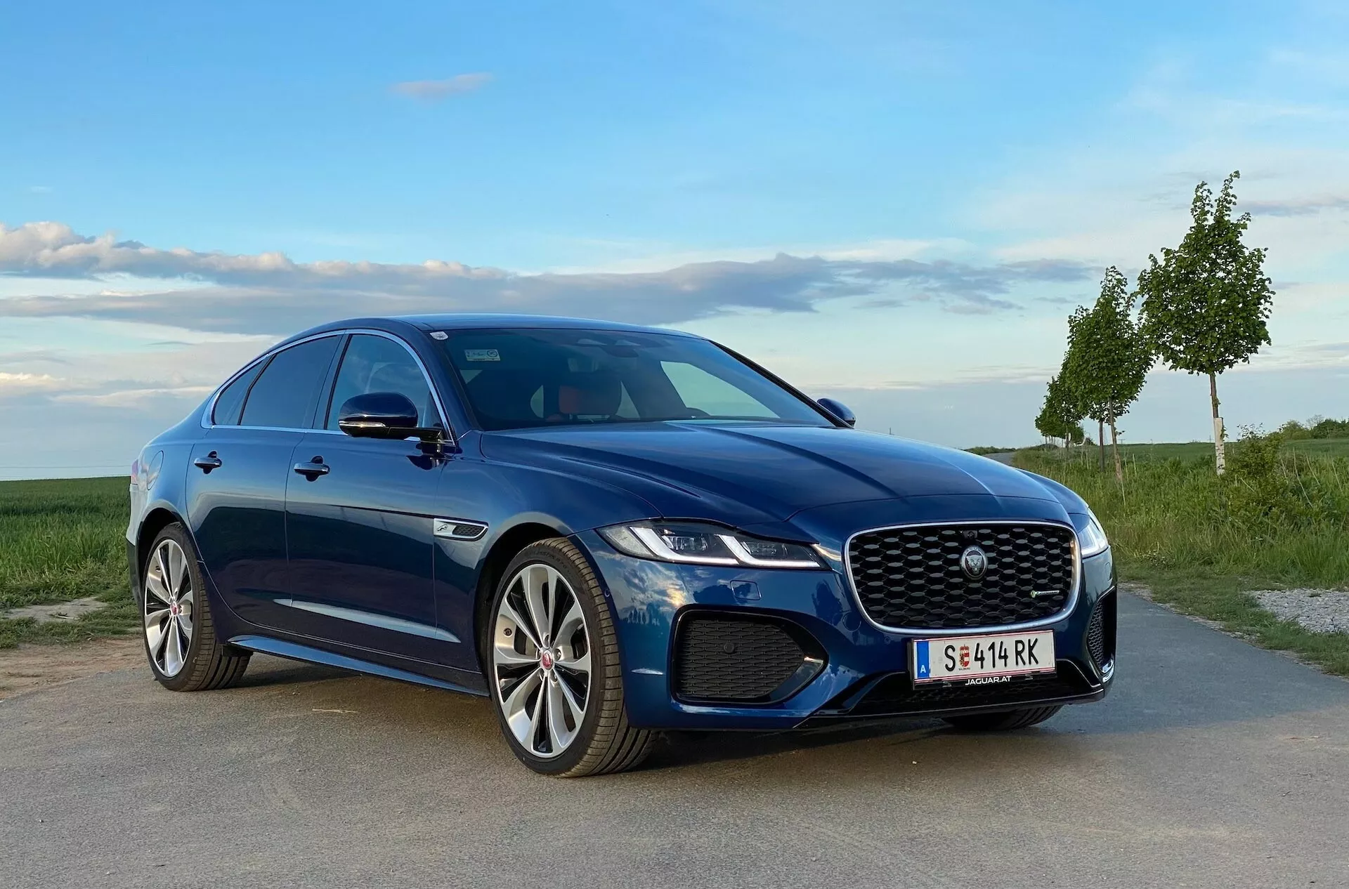 Jaguar XF 2021 blu, parcheggiata su una superficie pavimentata con un paesaggio erboso e un cielo chiaro sullo sfondo, mostrando una distintiva griglia, fari a LED, elegante paraurti anteriore con prese d'aria e cerchi in lega multispezzola. Il profilo sportivo della vettura è enfatizzato dalle linee eleganti e dalle minigonne laterali curve, vista da una prospettiva diagonale frontale a bassa angolazione.