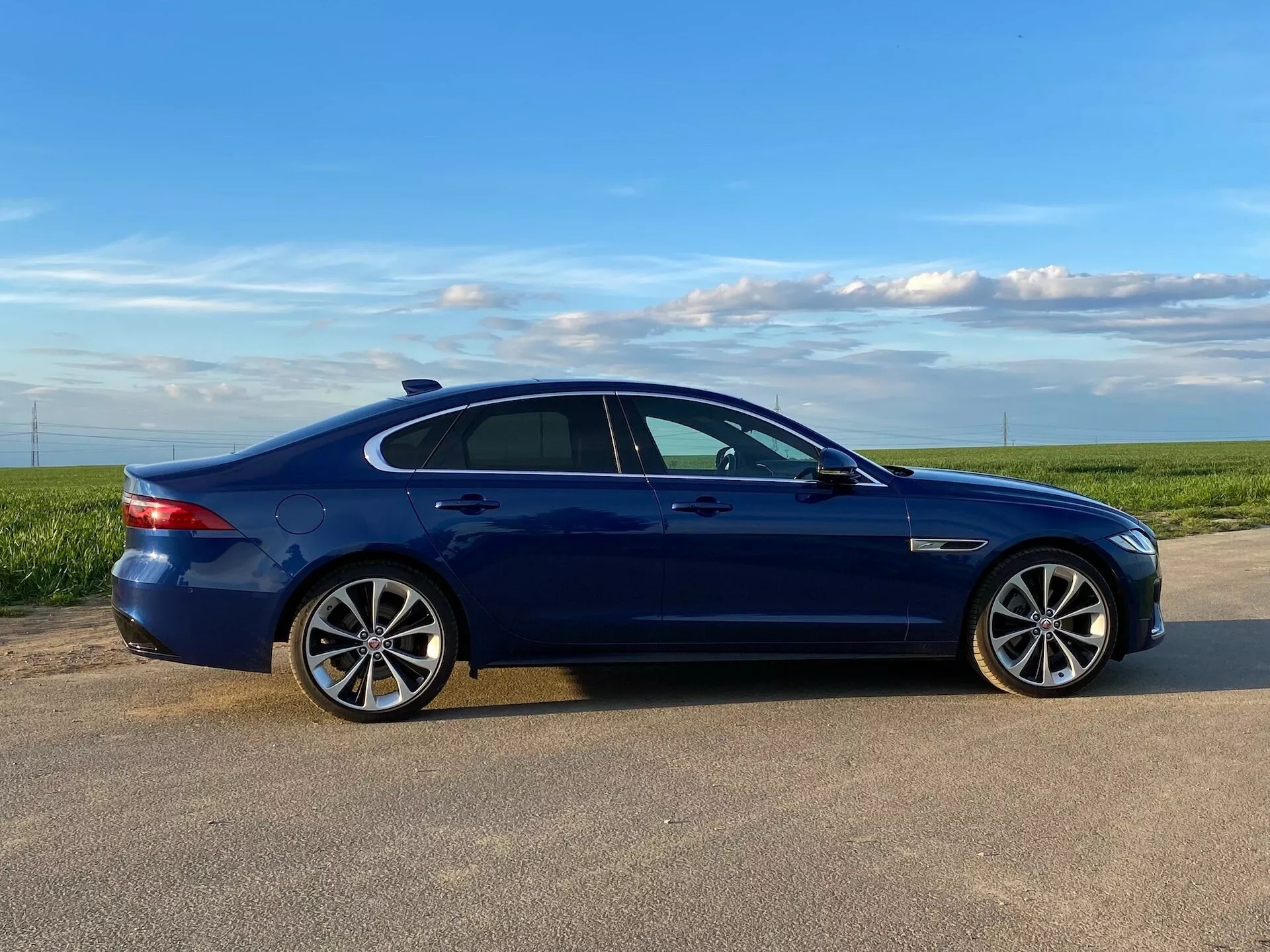 Una vista laterale del profilo della berlina Jaguar XF 2021 blu catturata alla luce del giorno, che mostra le sue caratteristiche di design come cerchi in lega multirazze, pneumatici a basso profilo, finestre oscurate, specchietti laterali del colore della carrozzeria con indicatori di direzione integrati, maniglie delle porte a filo del colore della carrozzeria e finiture cromate o in metallo spazzolato lungo la linea delle finestre. L'auto è posizionata su una superficie pavimentata con un campo aperto e un cielo chiaro sullo sfondo, che suggerisce un ambiente rurale o semi-rurale.